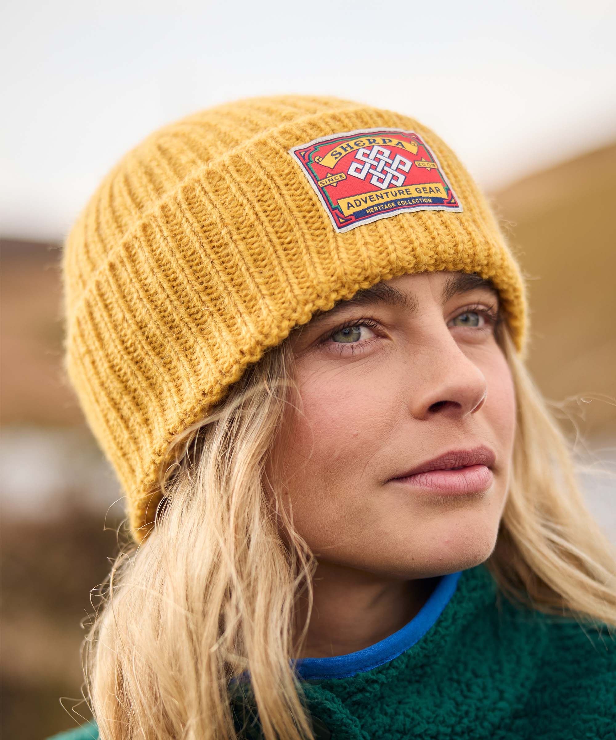 Close-up portrait of a woman standing outdoors on a hillside, looking ahead with a relaxed expression. Rolling hills and soft natural light create a scenic backdrop. She wears a mustard-yellow rib-knit beanie with a colorful woven patch and a teal fleece jacket.
