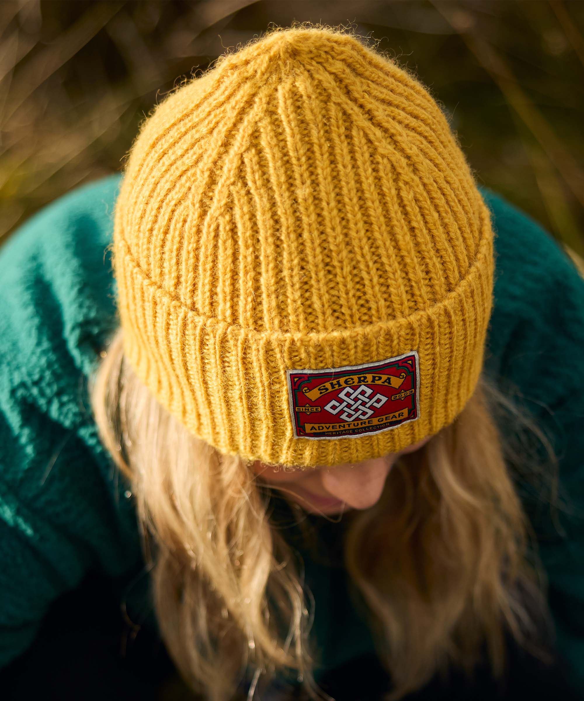 Woman leaning forward while outdoors, looking down toward the ground as if adjusting equipment or examining something nearby. The image is framed from above, highlighting the textured knit hat and natural surroundings. She wears a mustard-yellow rib-knit beanie with a woven patch and a teal fleece jacket.