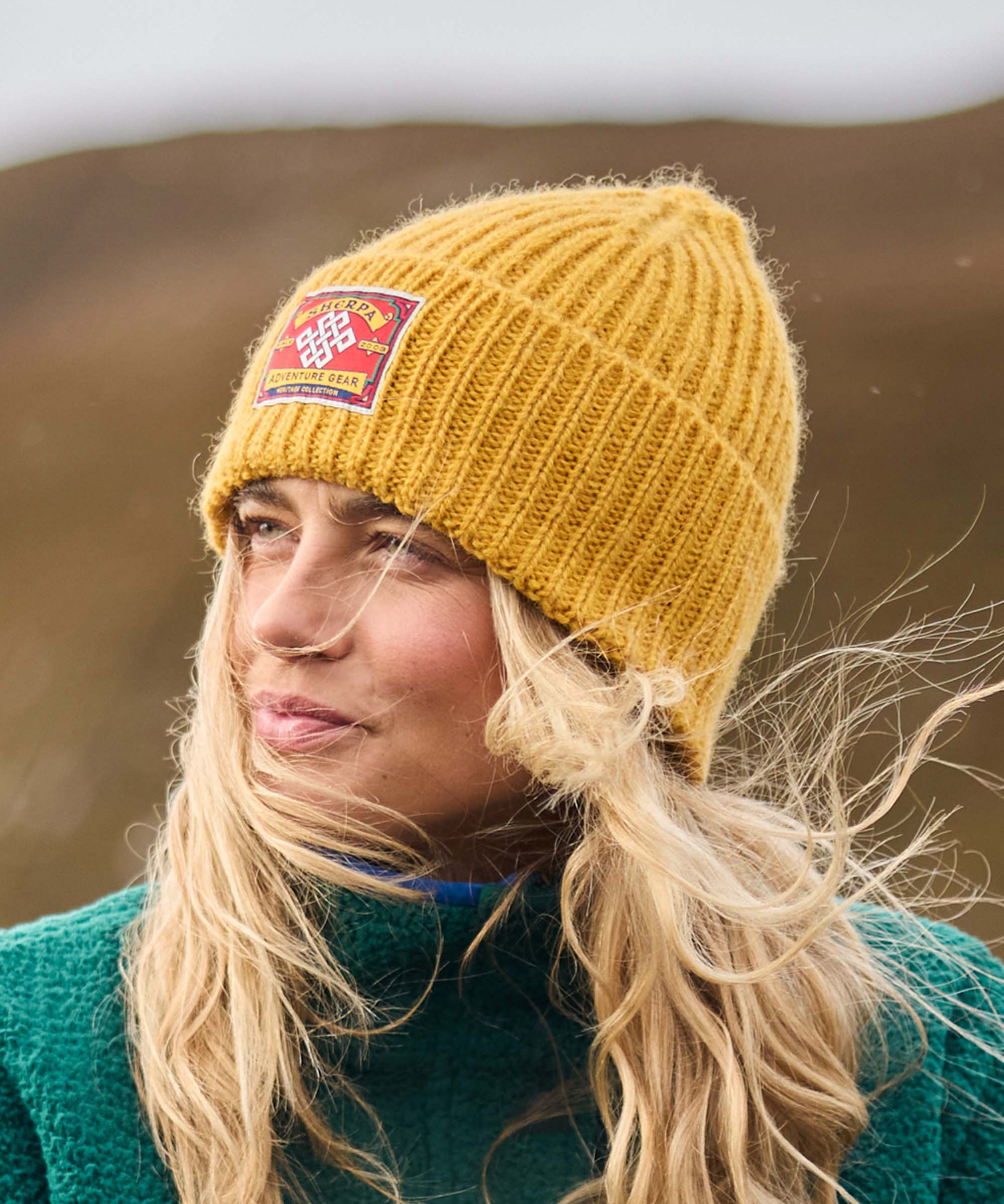 Woman standing in an open landscape with wind-blown hair, looking off into the distance with a slight smile. Hills and muted natural colors form the background. She wears a mustard-yellow rib-knit beanie with a woven patch and a teal fleece jacket.