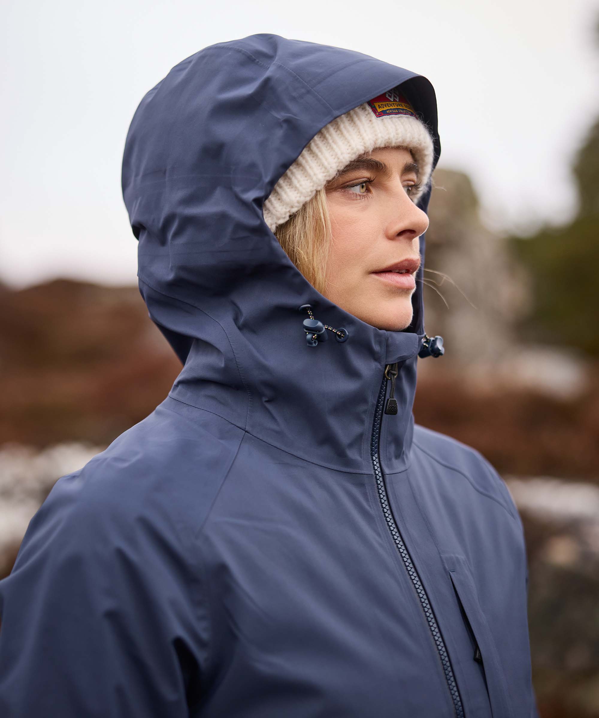 Close-up side profile of a woman standing outdoors with the hood of her blue waterproof jacket pulled up over a cream knit beanie. She gazes into the distance against a softly blurred mountain landscape, highlighting the jacket's streamlined hood and high collar.
