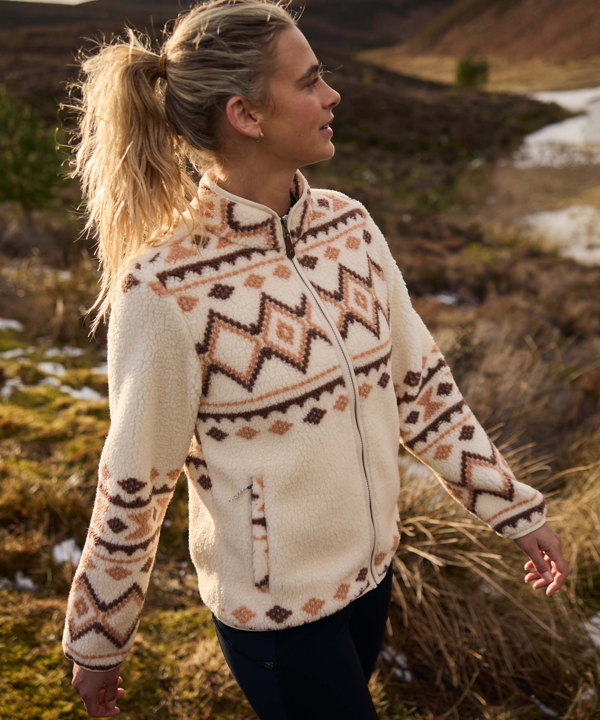 Woman walking through a grassy hillside landscape, turning her head to take in the scenery around her. The terrain is dotted with rocks, tall grasses, and patches of water. She wears a cream fleece jacket featuring a brown geometric Fair Isle-inspired pattern across the chest, sleeves, and shoulders, paired with dark leggings.