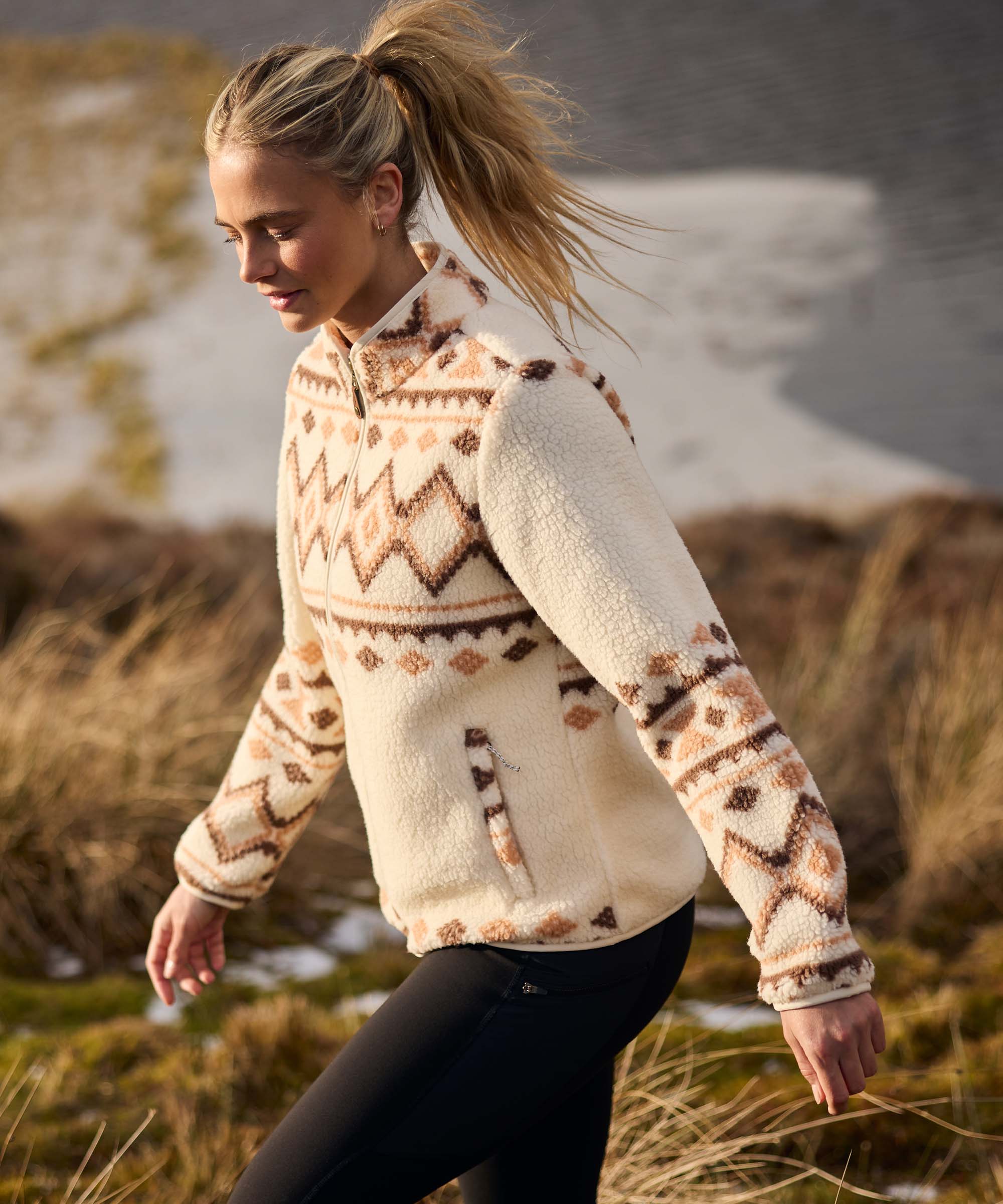 Woman striding across a rugged outdoor landscape with a relaxed smile, arms swinging naturally as she walks through tall grass. A lake and rolling hills are visible in the background. She wears a cream fleece jacket with an all-over geometric pattern in warm brown tones and black leggings.