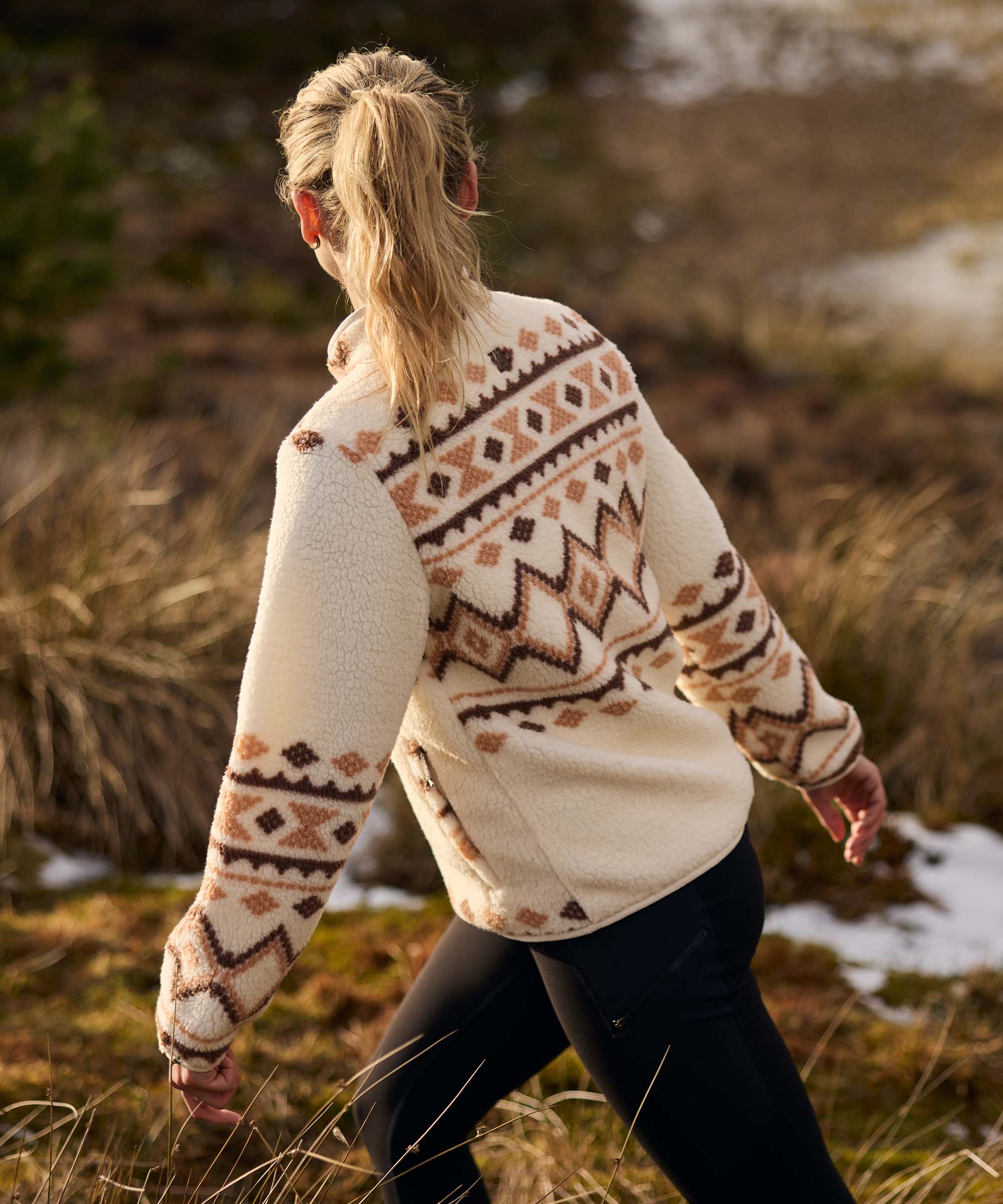 Rear view of a woman walking through a grassy hillside area near a body of water, exploring the landscape at an easy pace. Her blonde hair is tied back in a ponytail. She wears a cream fleece jacket decorated with a brown geometric knit-inspired pattern across the back and sleeves, paired with dark leggings.