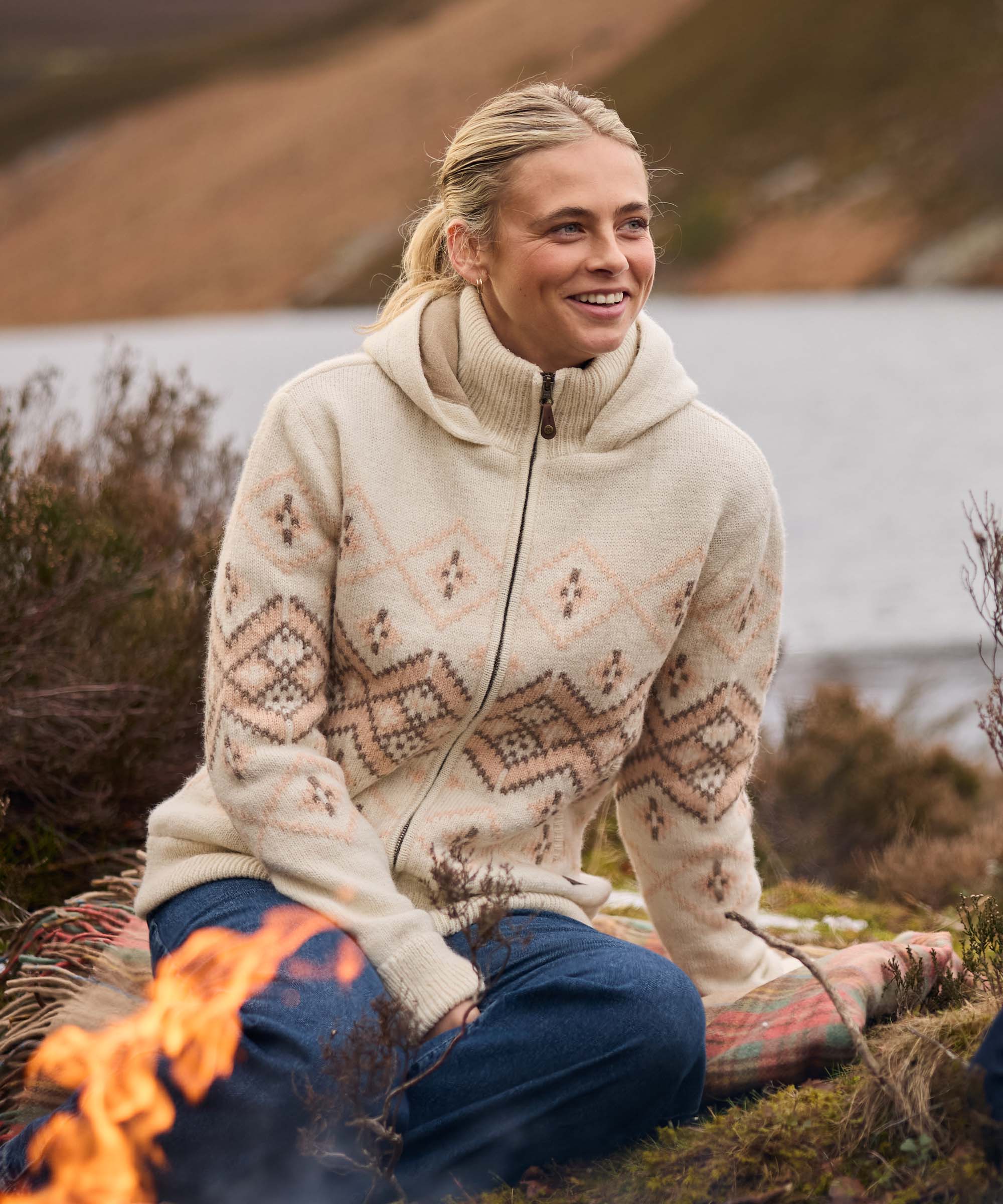 Woman sitting outdoors beside a small campfire, smiling while relaxing on a blanket near the waterÕs edge. The peaceful setting features a lake, rolling hills, and dry grasses. She wears a cream knit sweater with a soft geometric Fair Isle-inspired pattern in beige and blush tones, along with blue jeans.