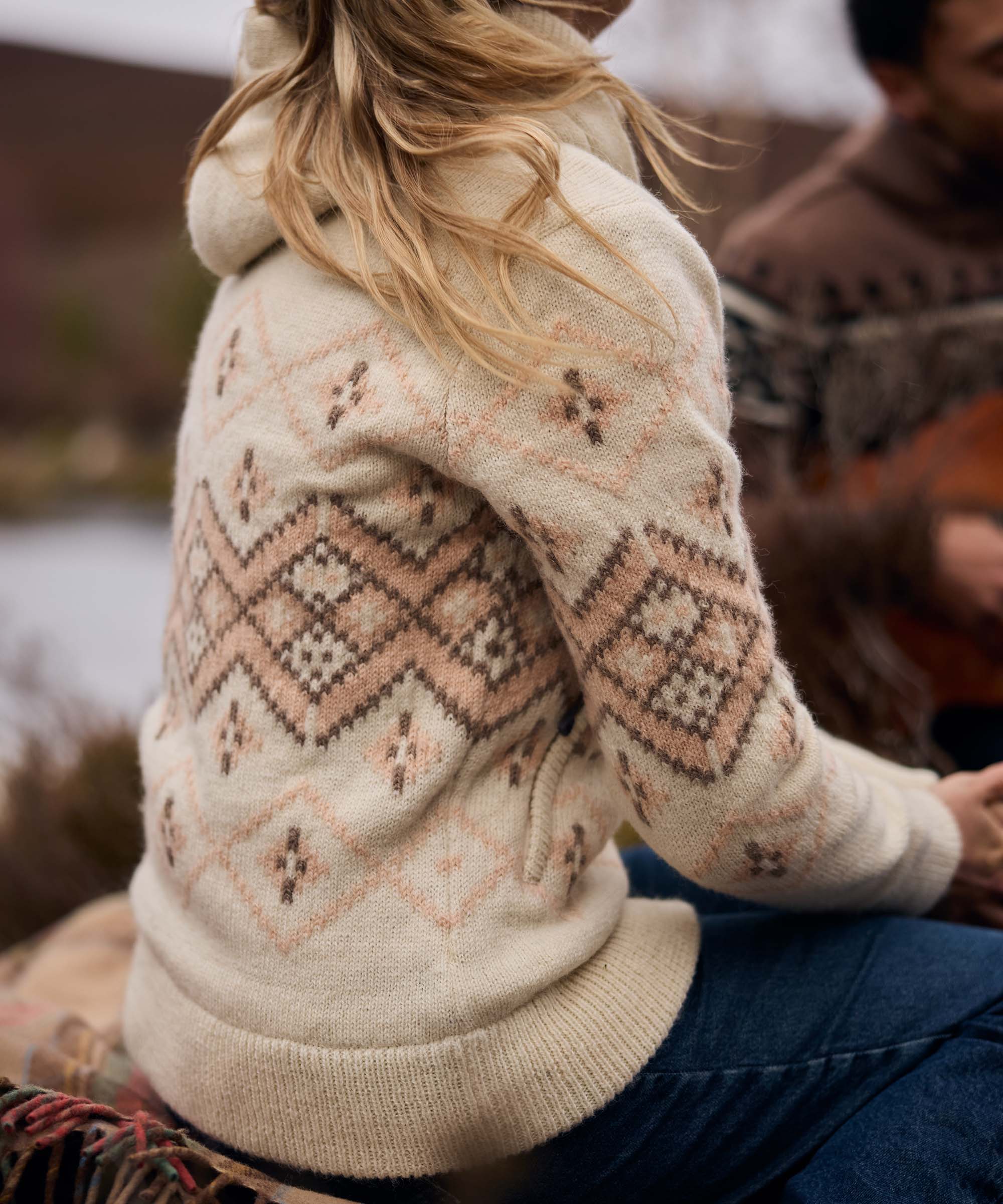 Woman seated outdoors beside a lake or river, turned slightly away from the camera while relaxing with companions in a rugged natural setting. The scene feels calm and social, with rocky terrain and muted autumn colors in the background. She wears a cream knit sweater featuring a geometric Fair Isle-inspired pattern in soft beige and blush tones, paired with blue jeans.
