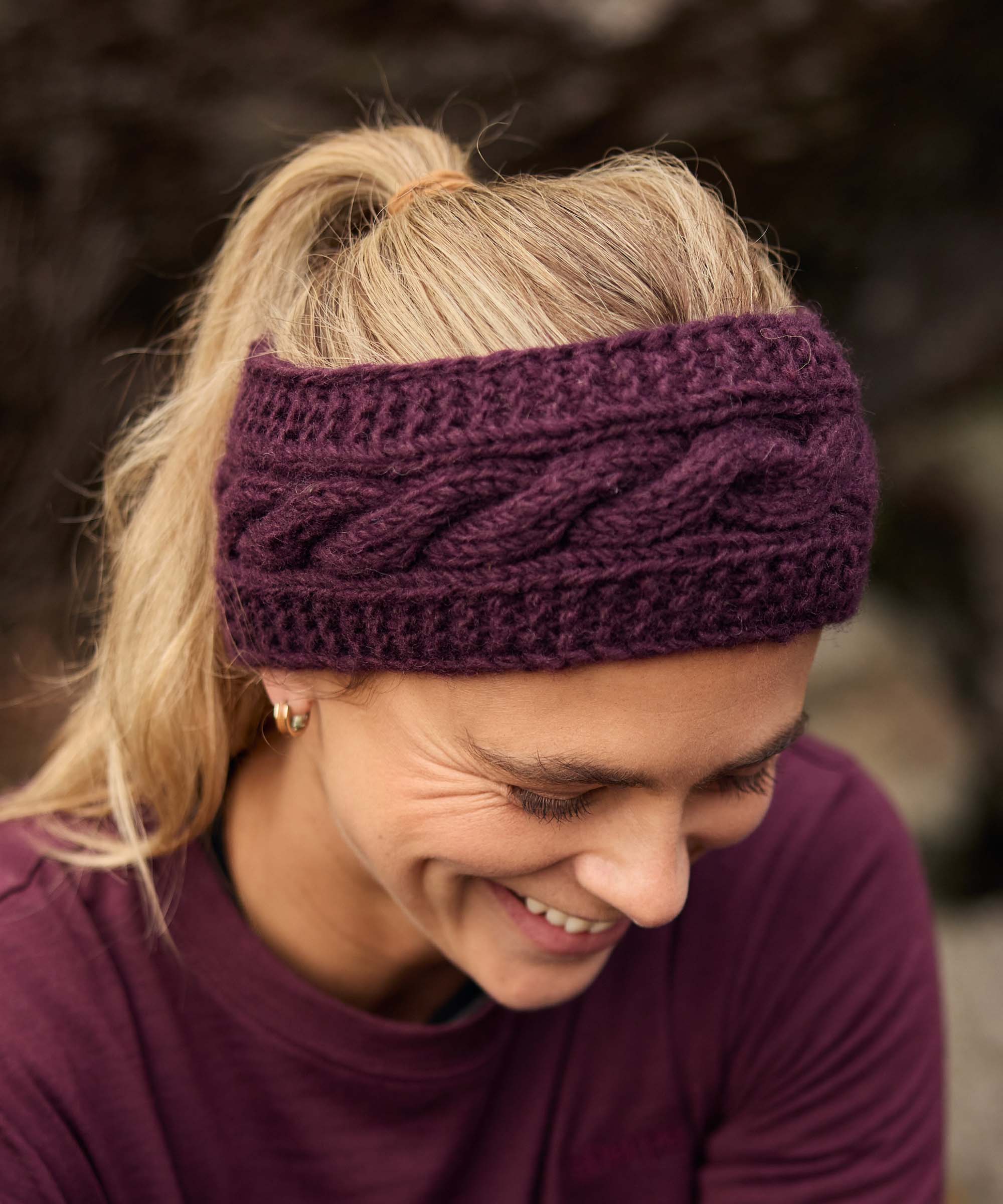 Woman sitting on rocks outdoors, looking down with a broad smile as she enjoys a quiet moment with friends. Natural stone formations and greenery create a relaxed mountain setting. She wears a plum-colored long-sleeve top and a wide cable-knit purple headband that holds her blonde hair back in a ponytail.