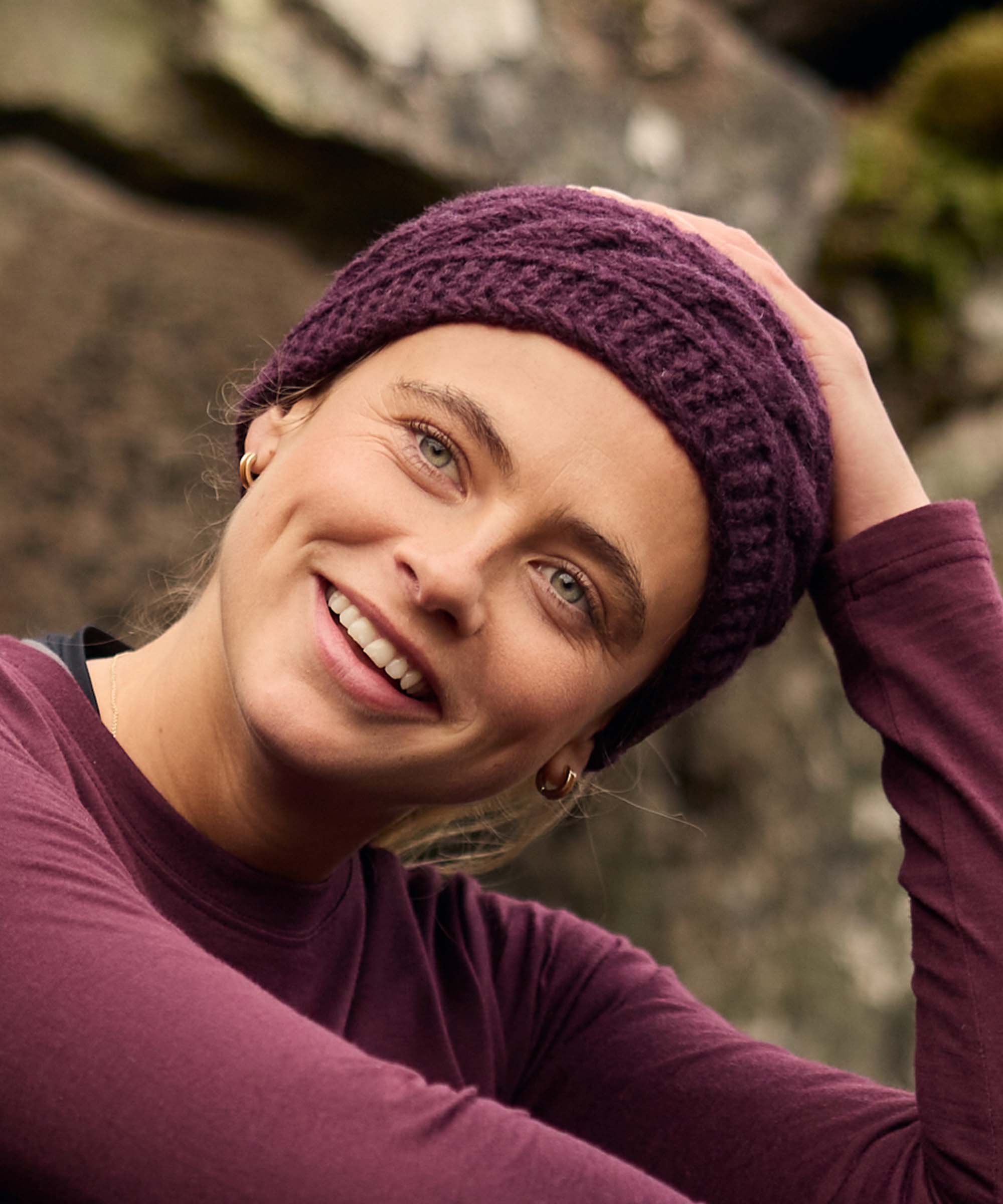 Woman seated among rocky terrain, smiling warmly toward the camera while resting her head against one hand. The outdoor setting features moss-covered rocks and soft natural light. She wears a purple cable-knit headband and a fitted plum long-sleeve top.