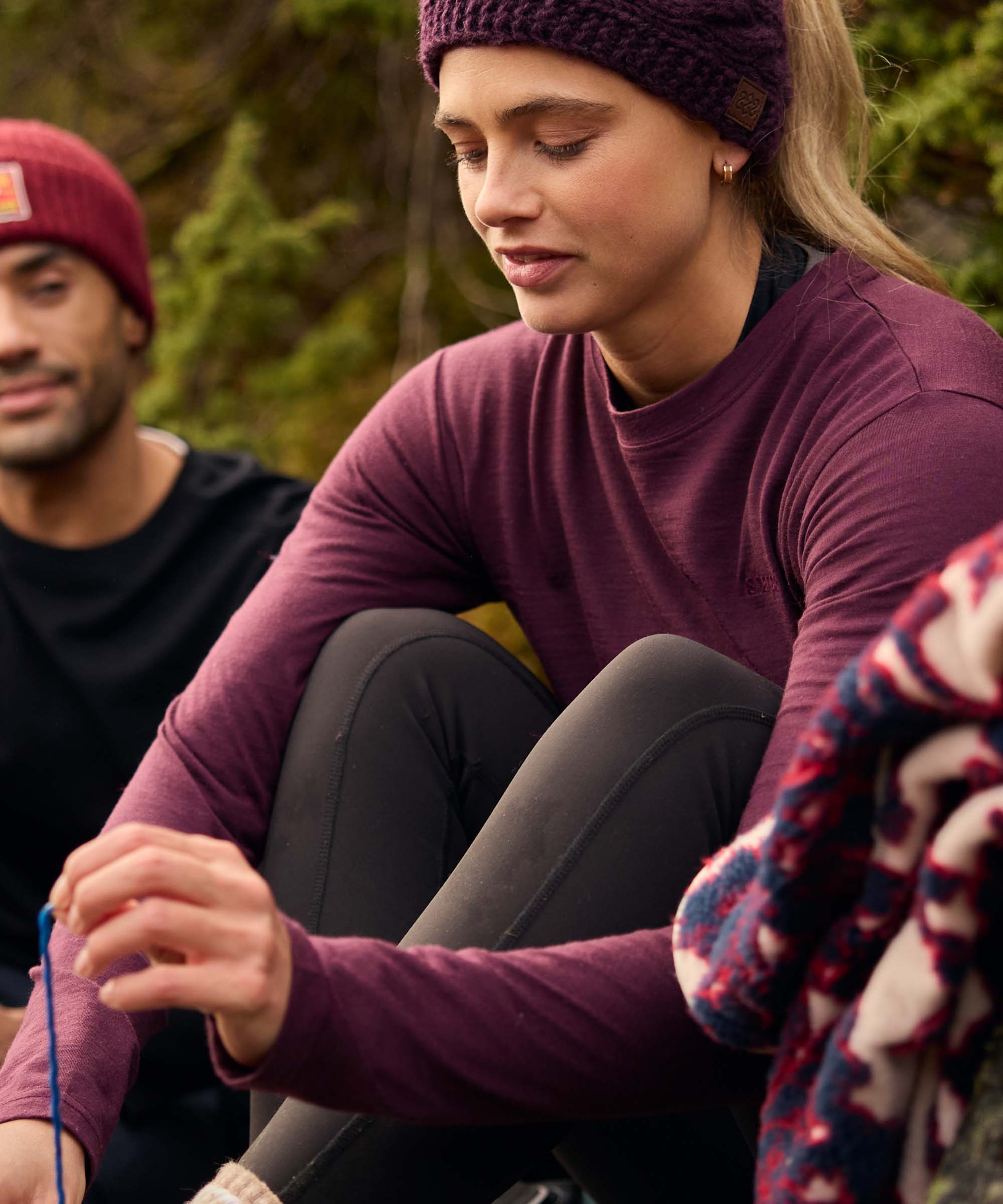 Woman sitting on rocks during an outdoor break, looking down as she reaches toward something in front of her while chatting with companions nearby. The rugged landscape and boulders suggest a hiking or climbing outing. She wears a plum-colored long-sleeve merino top, black leggings, and a purple knit headband.