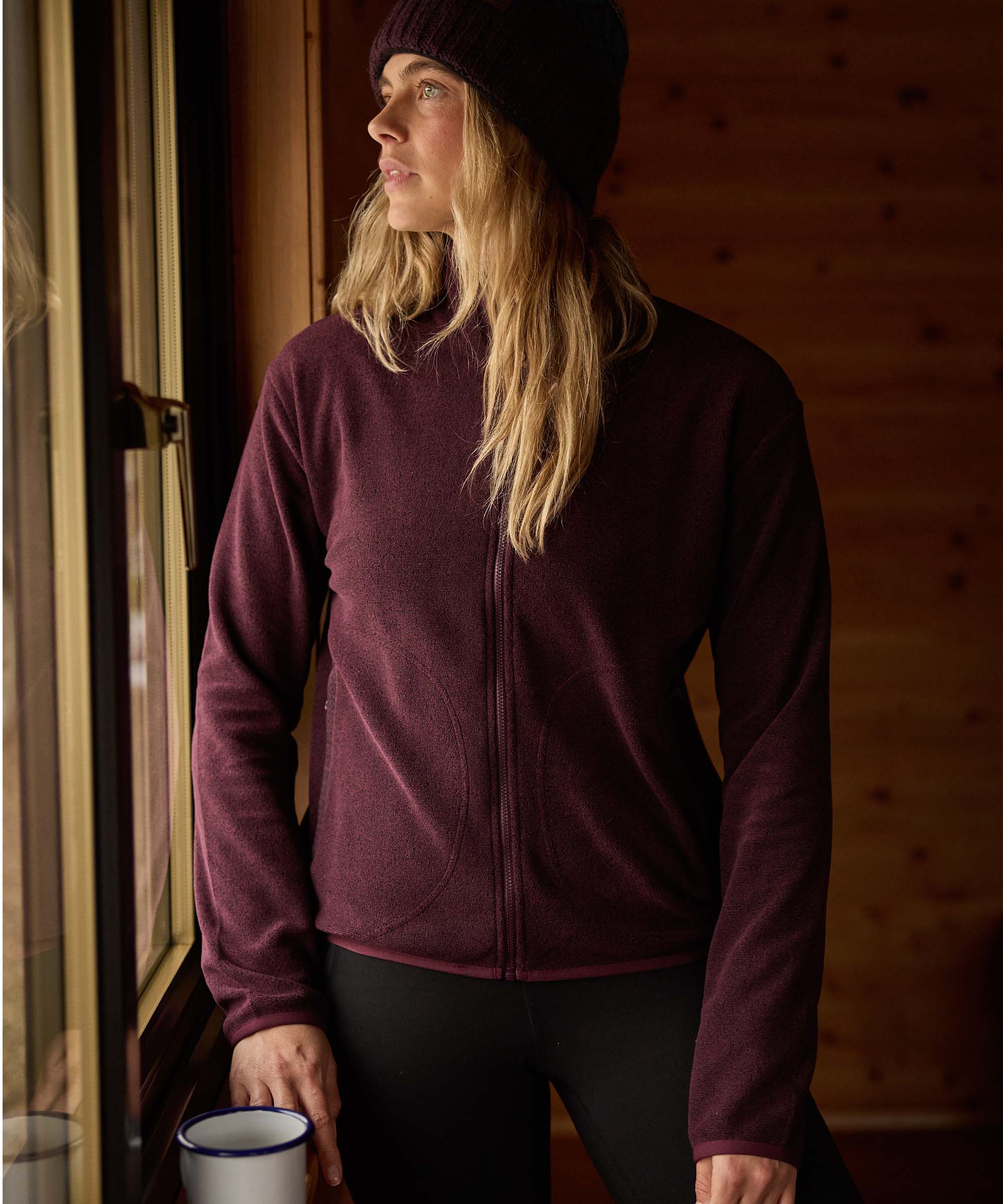 Woman standing beside a cabin window, gazing outside while holding a mug. Warm wooden walls and soft natural light create a cozy indoor atmosphere. She wears a burgundy full-zip fleece jacket, black leggings, and a dark knit beanie.