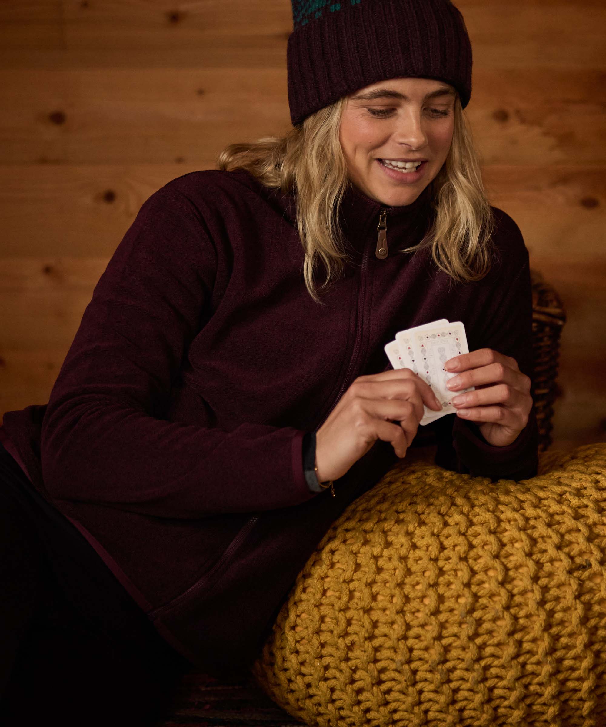 Woman relaxing indoors on a sofa or cushioned seat, smiling while holding a deck of playing cards. The wooden cabin interior and knitted blanket create a warm, comfortable setting. She wears a burgundy fleece jacket and a matching dark knit beanie.