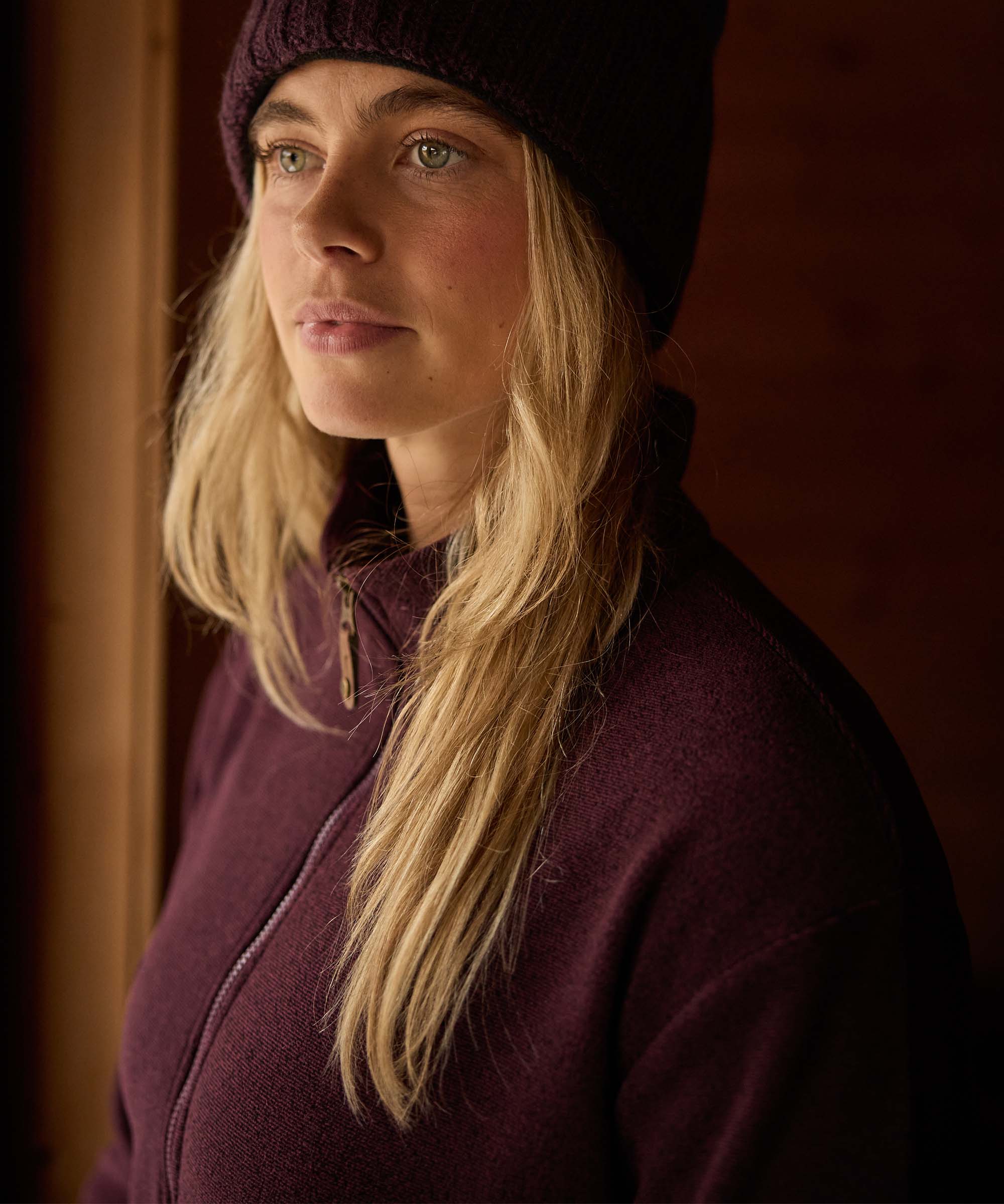 Close-up portrait of a woman standing inside a rustic cabin, looking thoughtfully out of frame toward a nearby window. Warm wood-paneled walls form the background. She wears a burgundy full-zip fleece jacket and a dark knit beanie, with her blonde hair falling over one shoulder.
