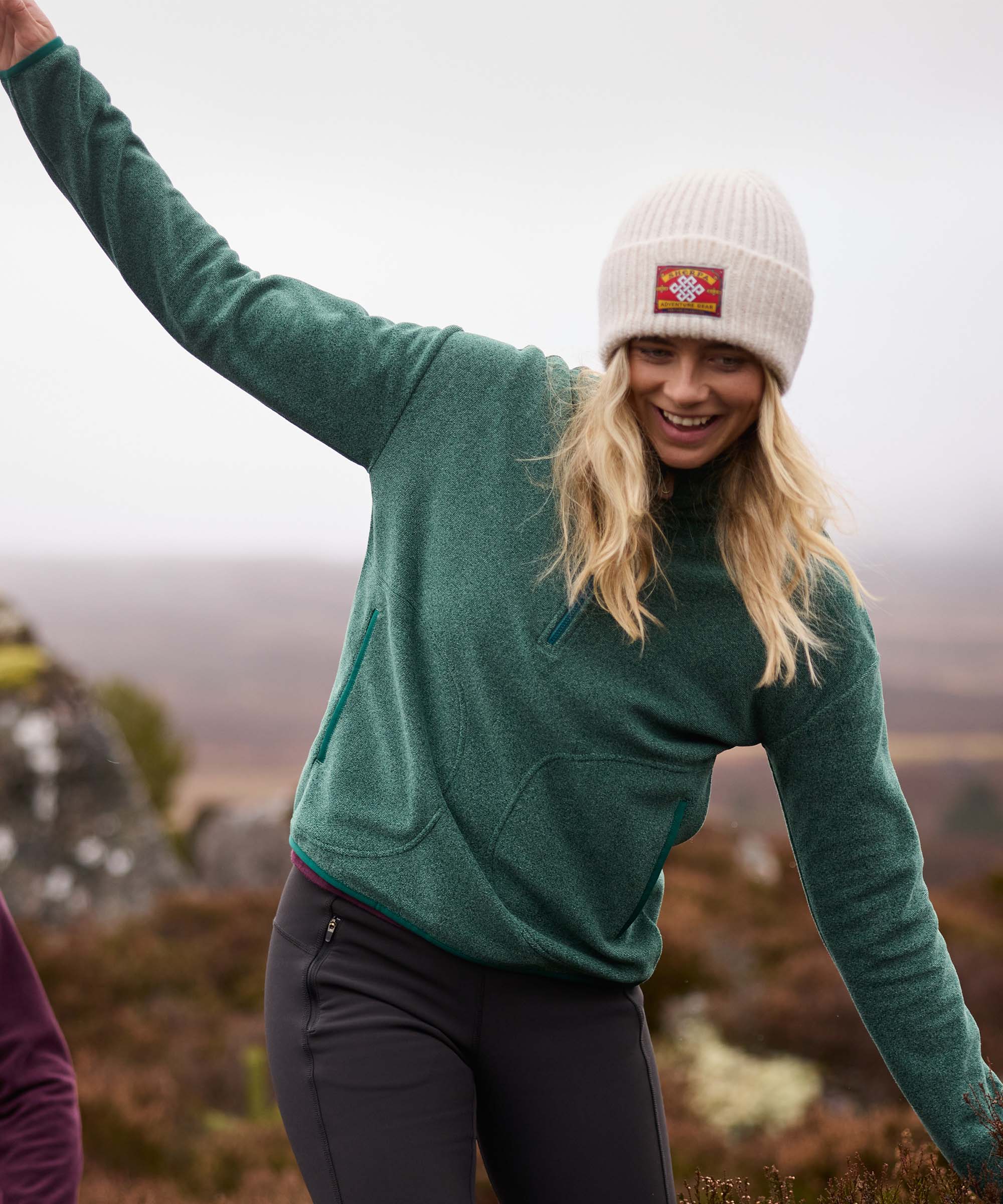 Woman balancing with her arms extended while stepping across uneven rocks on a hillside trail. The overcast landscape stretches behind her as she smiles and looks downward. She wears a green half-zip fleece pullover, black leggings, and a cream knit beanie.