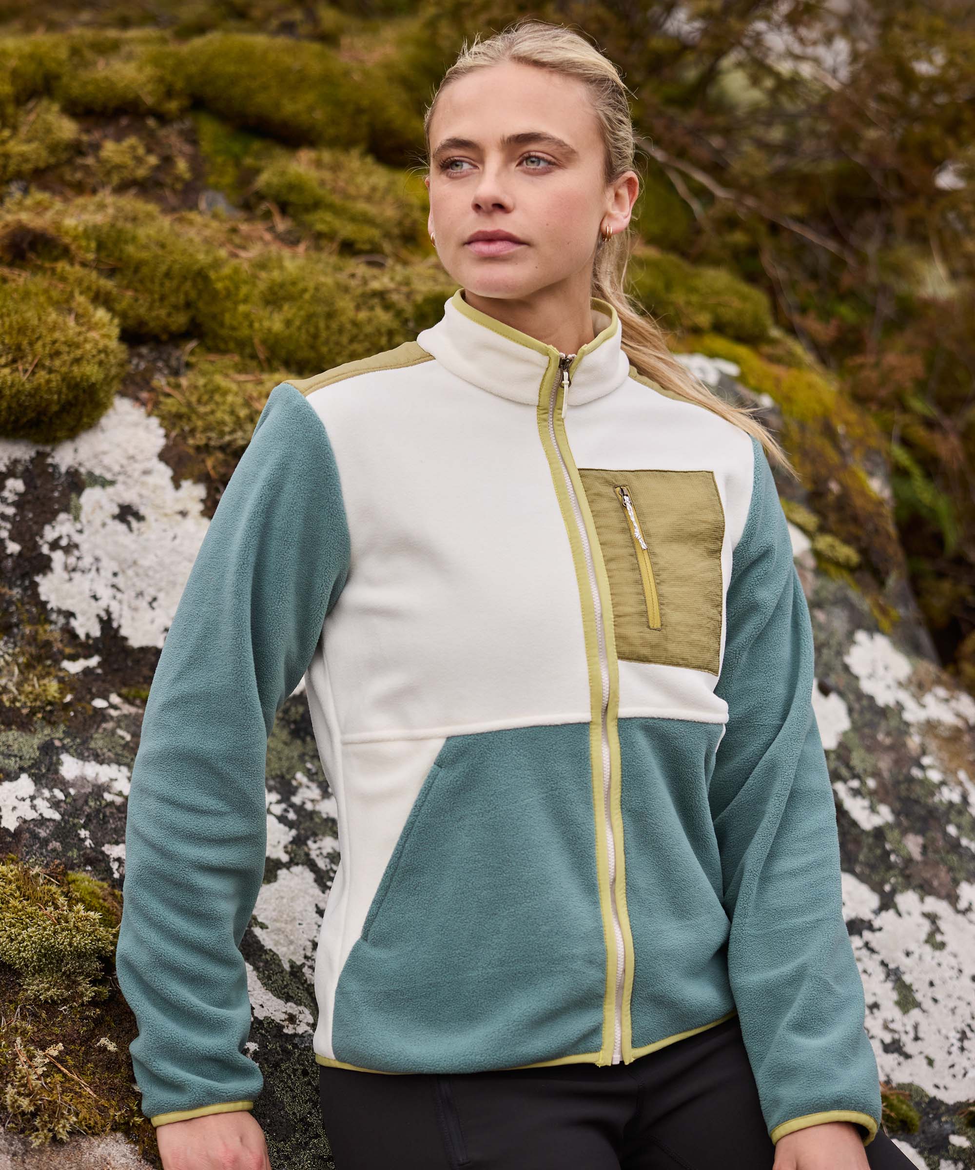 Woman standing beside a large moss-covered rock, pausing to look across the surrounding landscape. She appears relaxed and observant in an outdoor setting with natural rock formations and vegetation. She wears a color-block fleece jacket with teal sleeves, cream panels, and a zip chest pocket.
