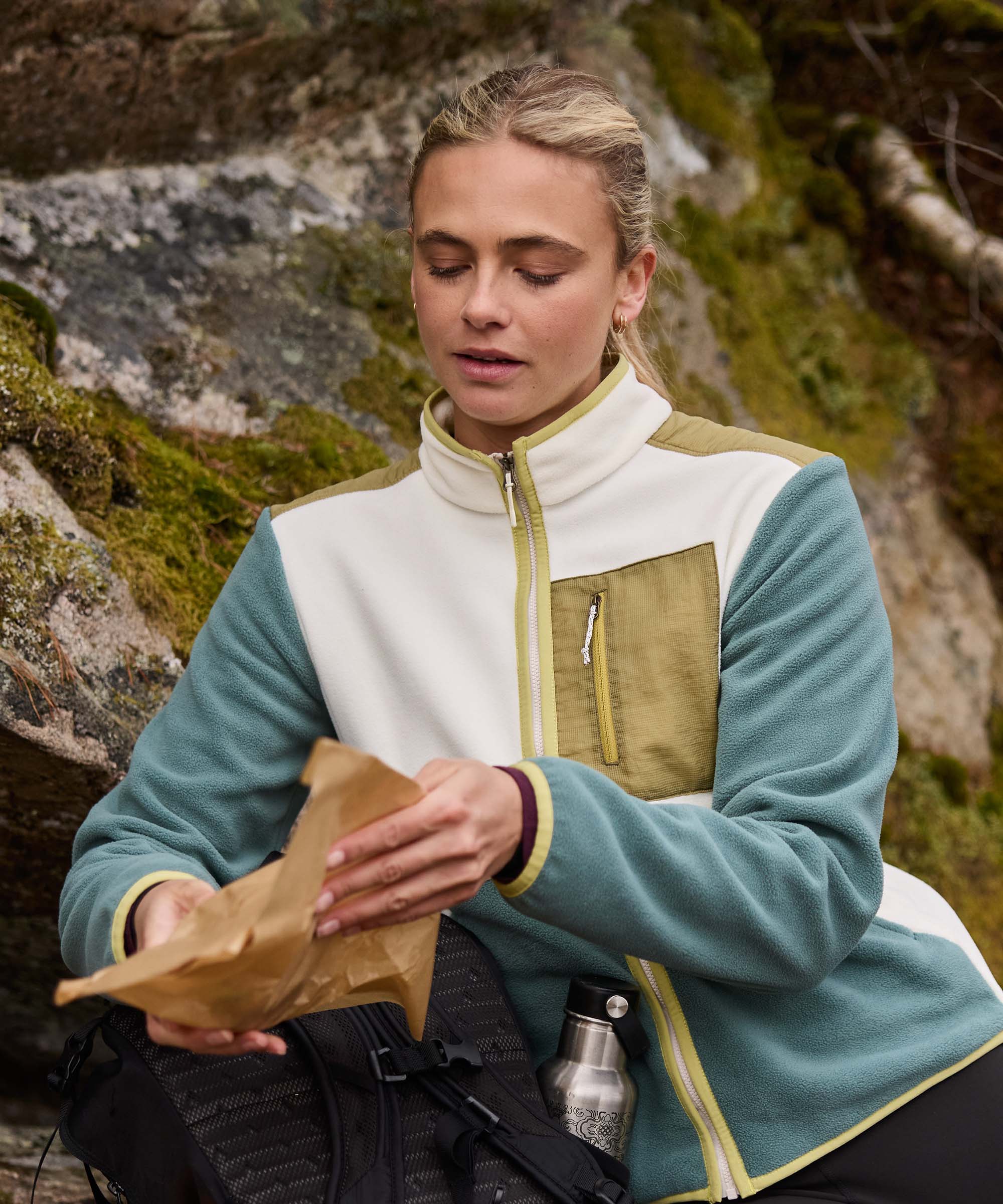 Woman seated on a rocky outcrop during a trail break, looking down as she reaches into a small paper bag. Mossy rocks and rugged terrain surround her. She wears a teal and cream color-block fleece jacket with contrasting trim and black leggings.