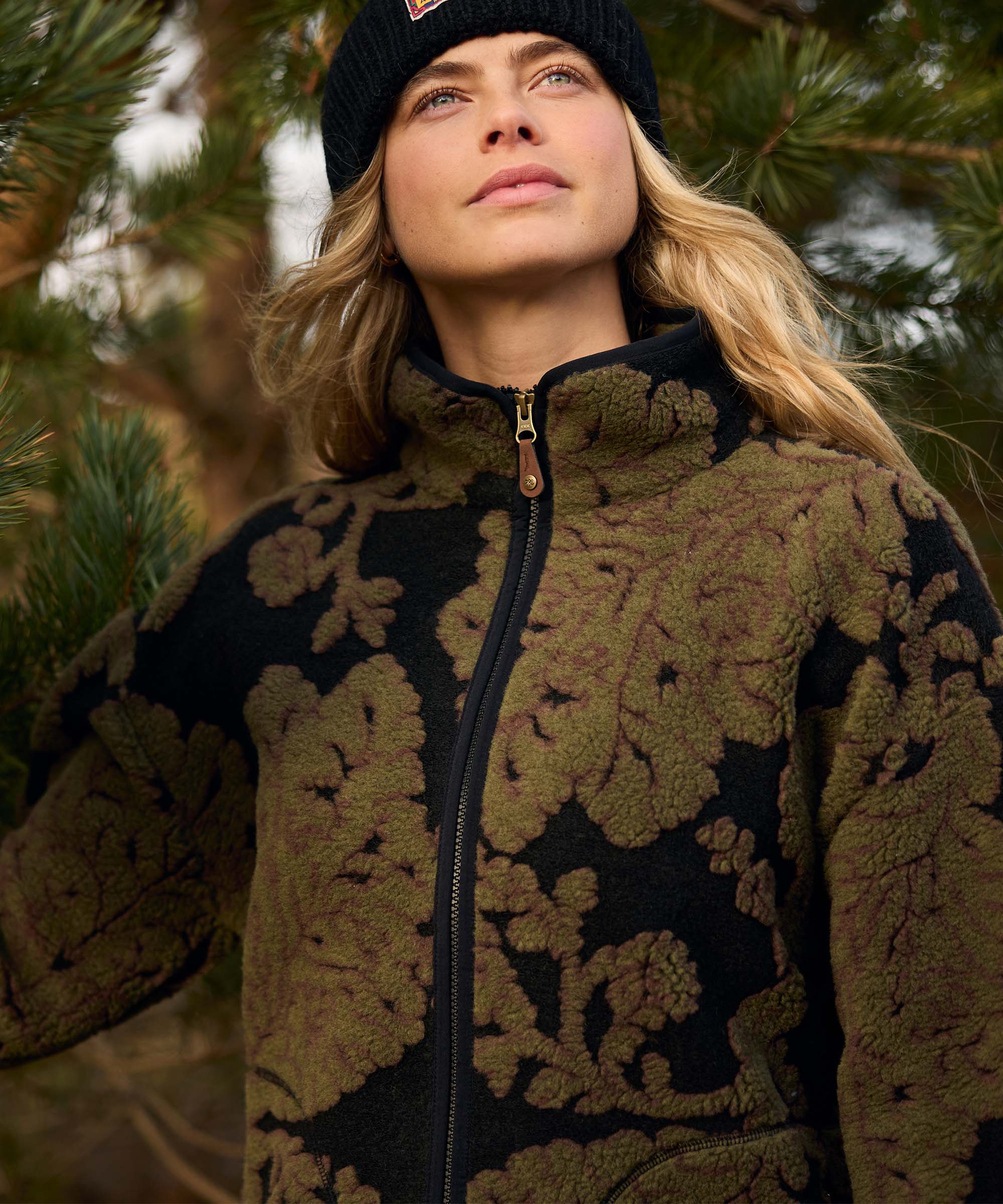 A woman stands beneath tall pine trees, looking upward with a calm expression. She wears a black knit beanie and an olive-green full-zip fleece jacket featuring a bold oversized floral pattern in contrasting textured fabric. The woodland backdrop complements the nature-inspired design.