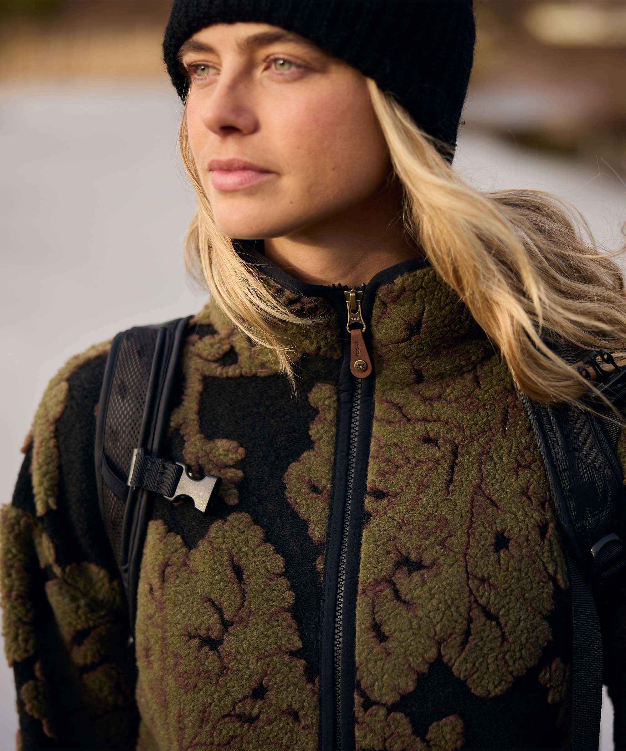 Close-up portrait of a woman wearing a black knit beanie and an olive-green floral-patterned full-zip fleece jacket. Backpack straps rest over her shoulders as she looks into the distance, suggesting she is preparing to continue her hike through the surrounding countryside.