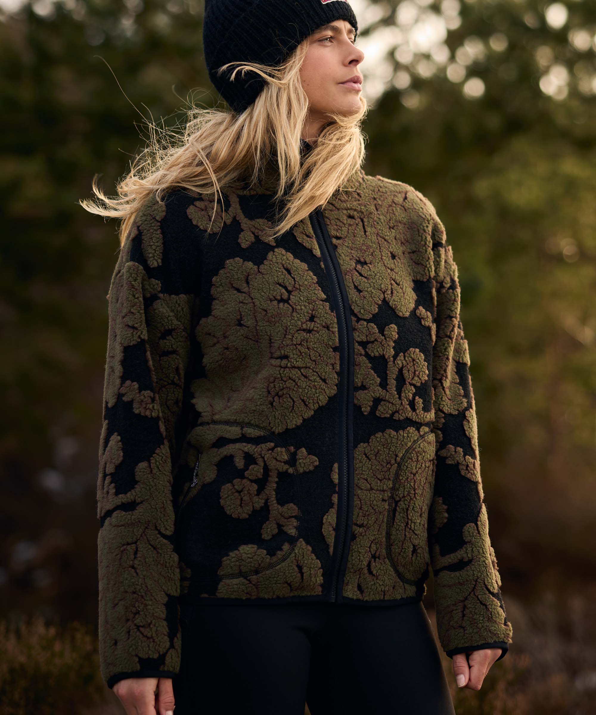 A woman stands outdoors surrounded by trees, turning her head to admire the scenery. She wears a black knit beanie and an olive-green fleece jacket with an oversized floral jacquard pattern and full-length zip. The relaxed pose captures a quiet moment in nature.