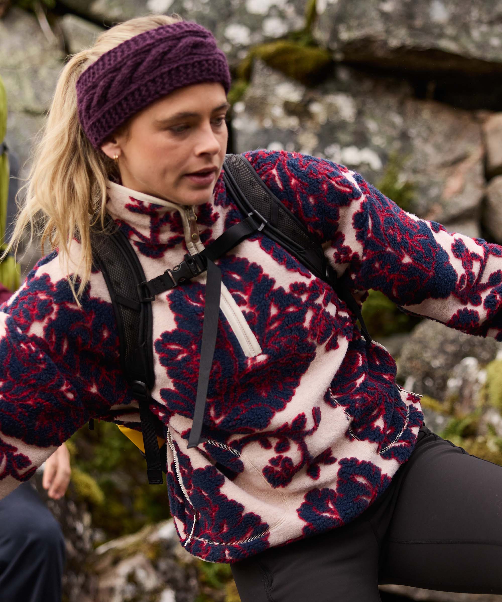 A woman navigates rocky terrain during a climbing or hiking excursion, extending her arm for balance as she moves across boulders. She wears a plum cable-knit headband and a quarter-zip fleece pullover with a bold floral print in deep red and cream tones.