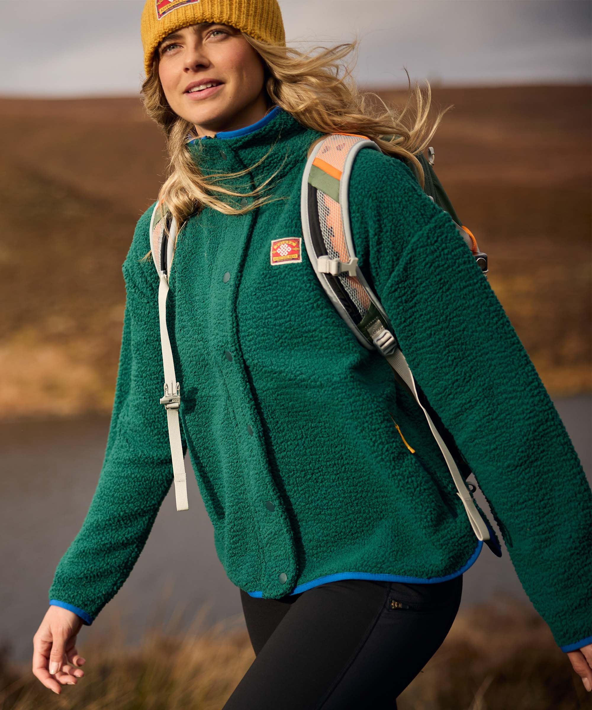 A woman hikes through a rugged landscape with a backpack over her shoulders, captured mid-stride during an outdoor excursion. She wears a green sherpa-style fleece jacket with a full-zip front and high collar, paired with a mustard knit beanie.