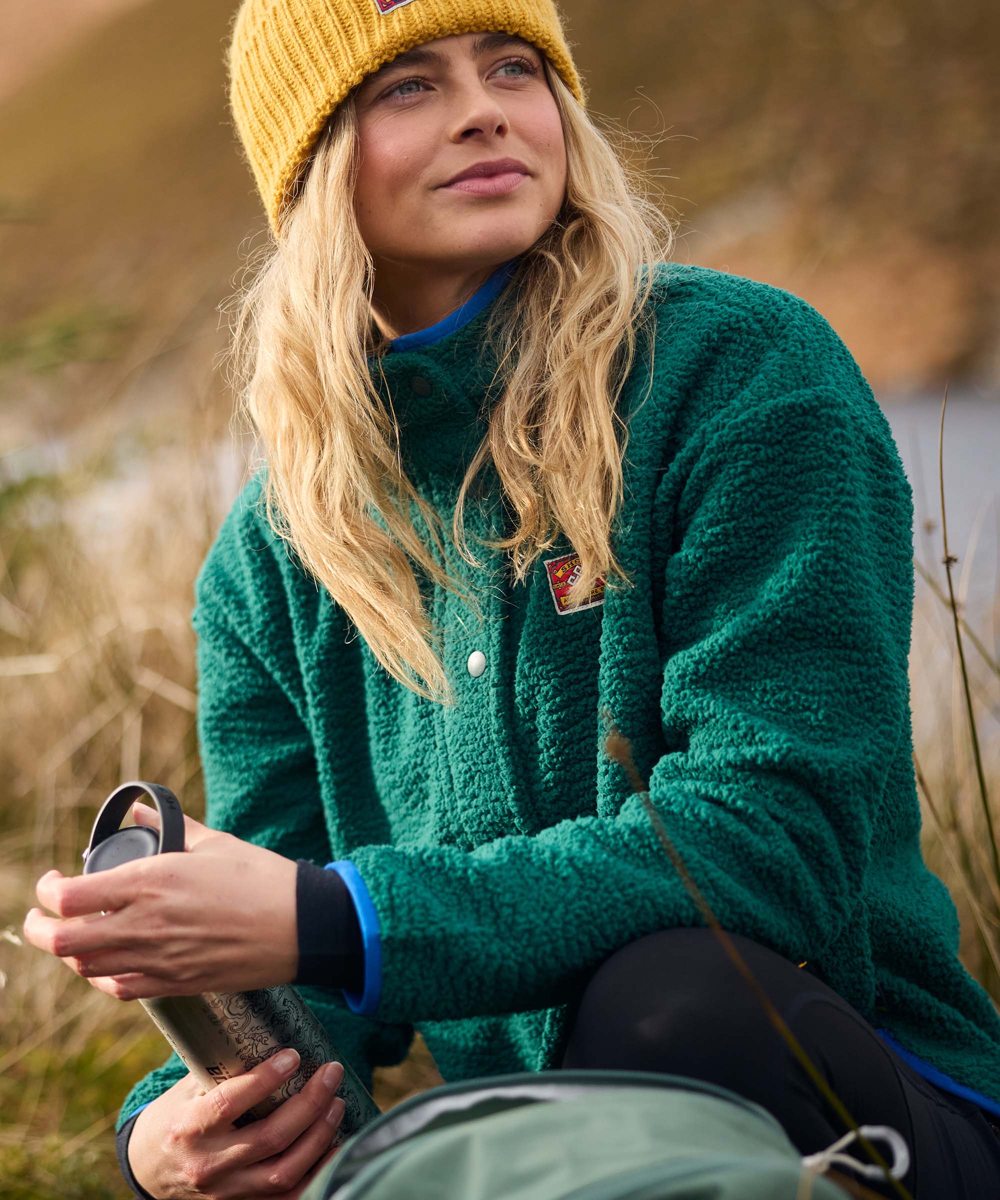 Taking a break beside her backpack, a woman sits outdoors and looks upward toward the surrounding scenery. She wears a textured green full-zip fleece jacket with a stand collar and a mustard yellow rib-knit beanie, creating a cozy cold-weather outfit.