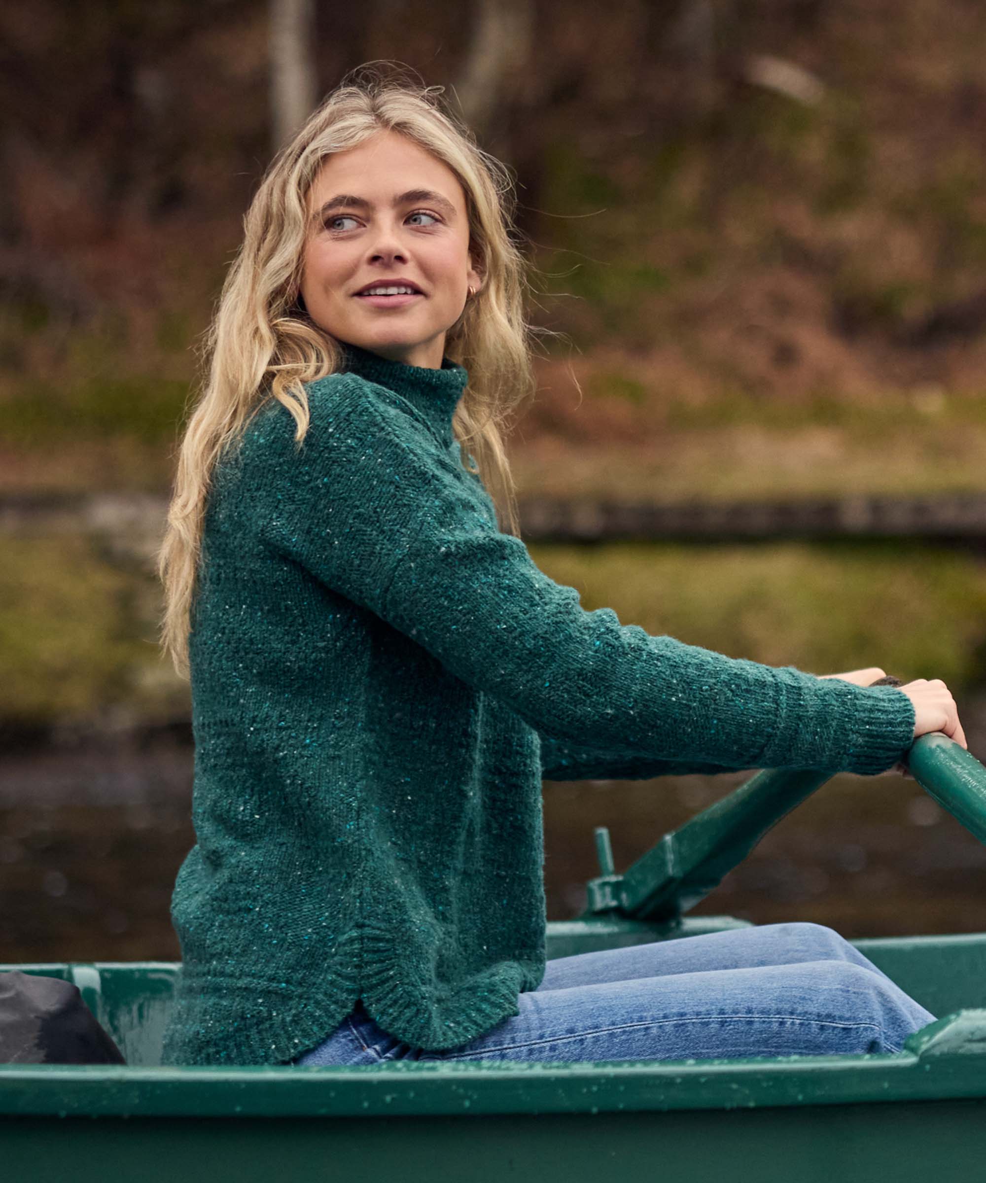 A woman rows a small green boat across calm water, turning to look behind her while enjoying time outdoors. She wears a forest green turtleneck sweater with a soft knit texture, long sleeves, and a relaxed fit suitable for cool-weather layering.