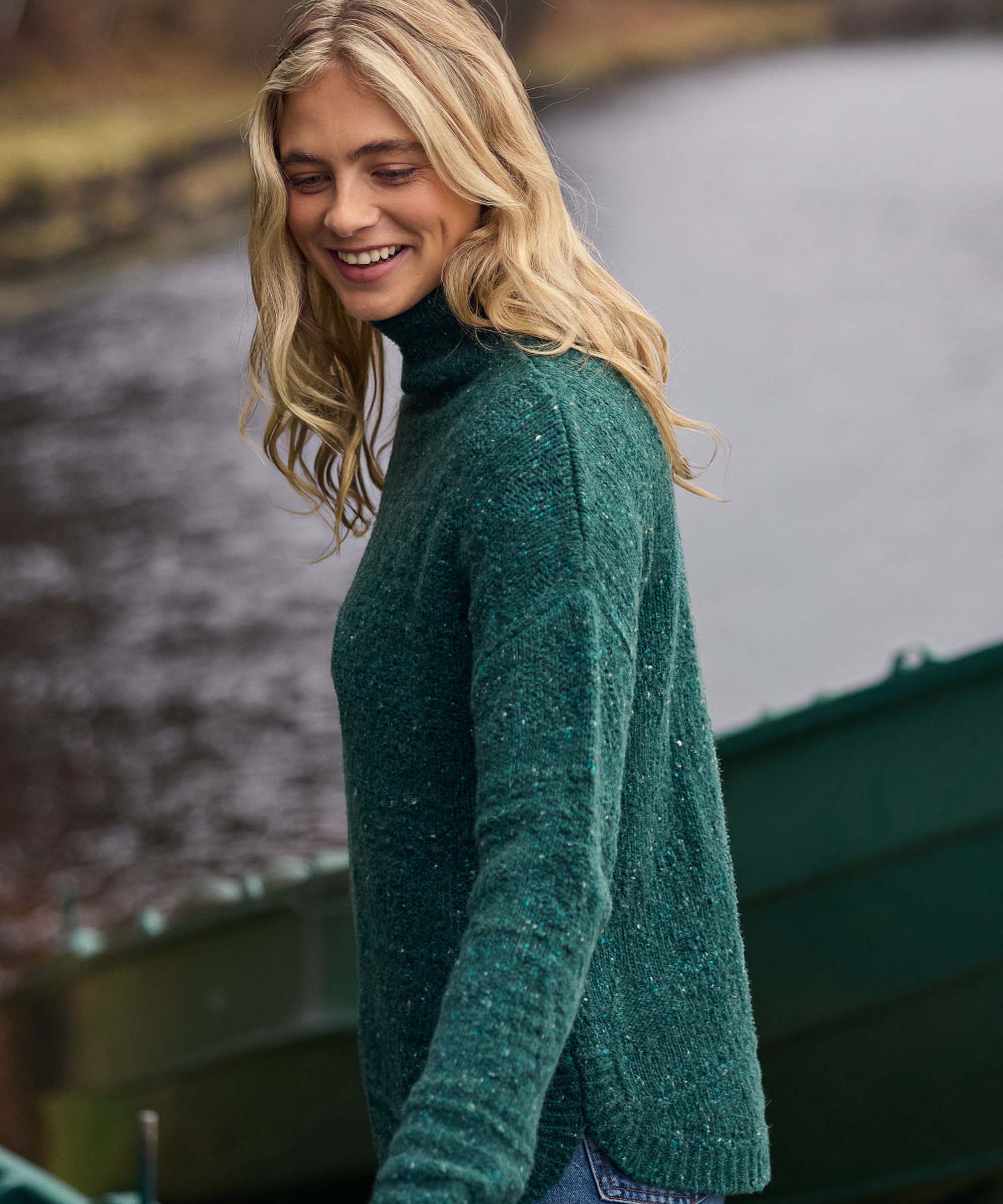 Standing in a small boat on a quiet lake or river, a woman smiles while taking in the surrounding scenery. She wears a green turtleneck knit sweater featuring a soft heathered texture, long sleeves, and a comfortable classic fit.