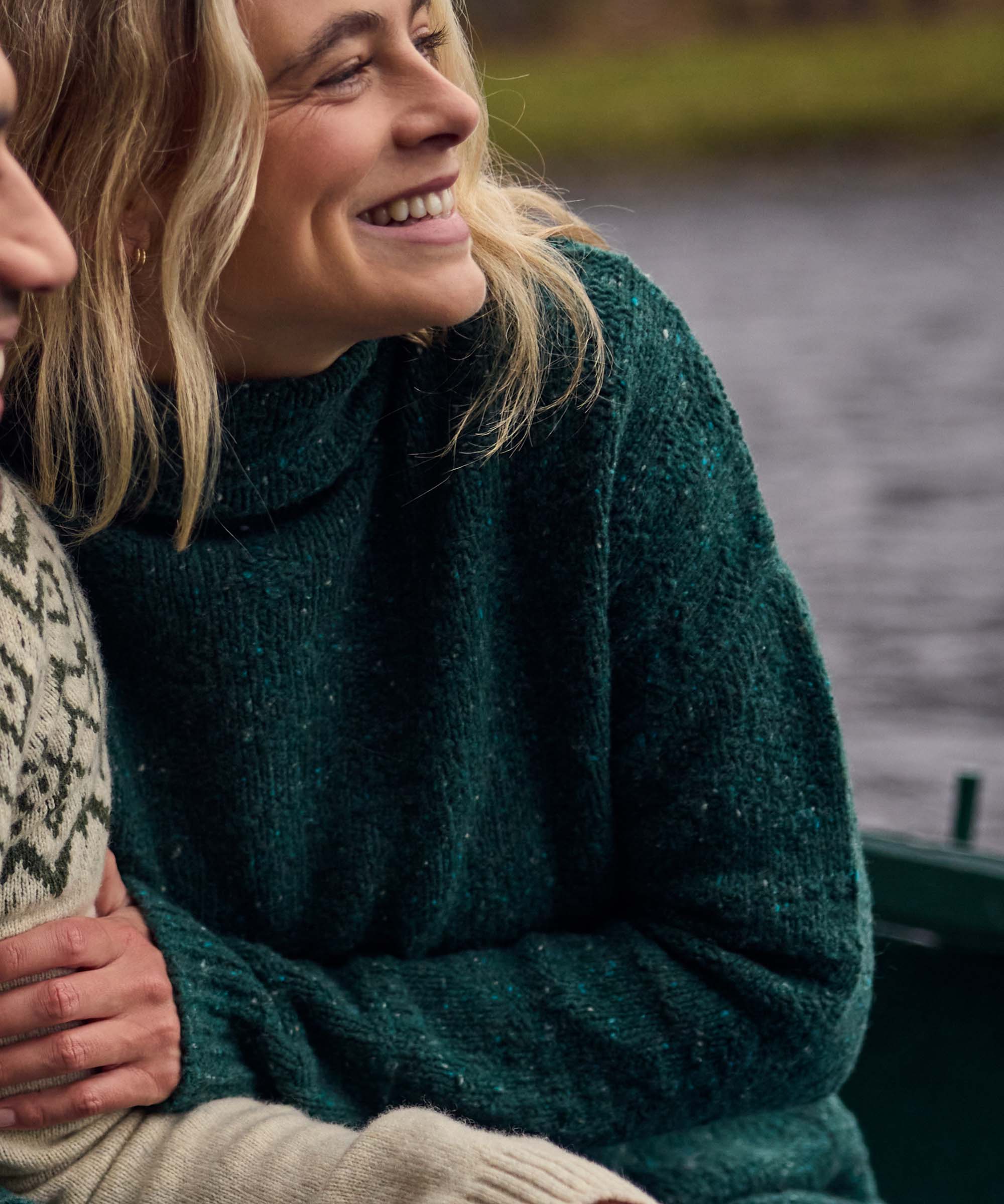 A close up of a woman sitting in a boat with a lake and green shoreline visible in the background. She is wearing a knitted emerald sweater with a subtle texture. The woman is holding the arm of another passenger.