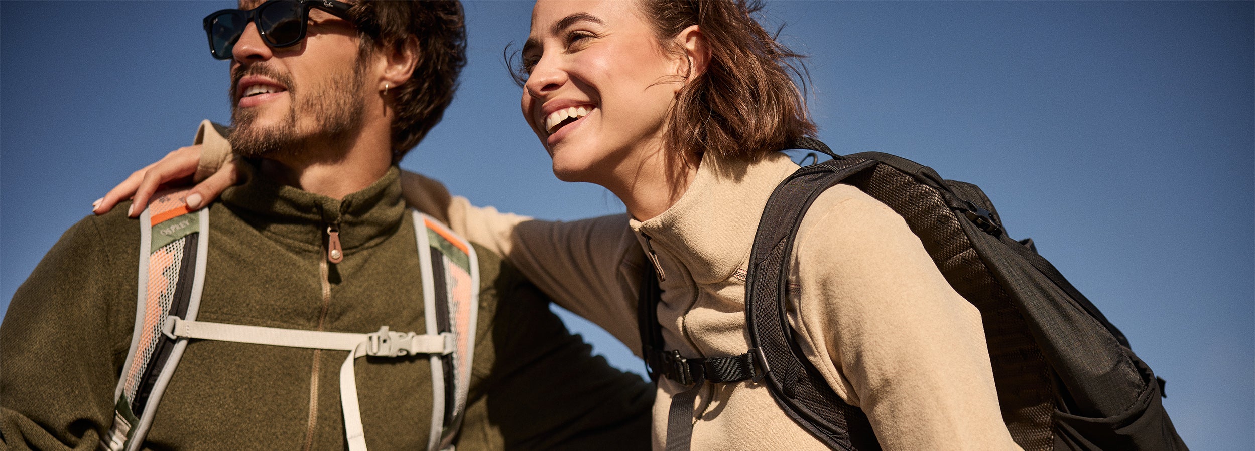 Two people with backpacks standing close together outdoors on a clear day. The man is wearing a green half zip fleece with cream
