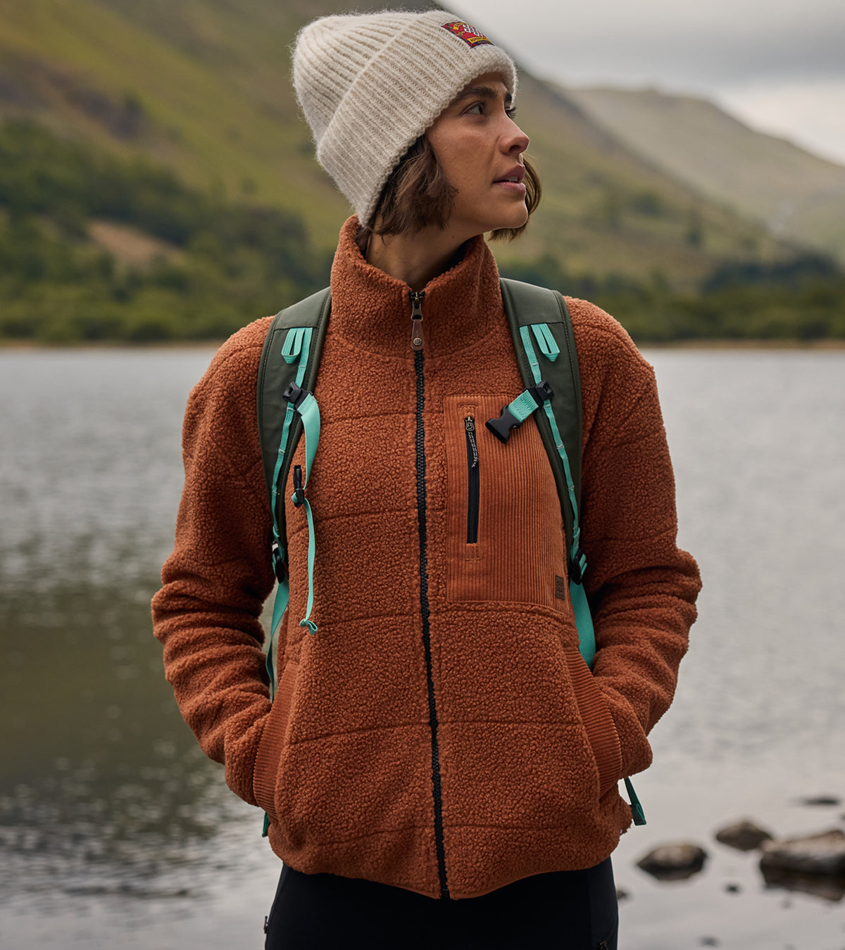A women is standing by a lake wearing brown Sherpa Adventure fleece jacket and a beige beanie. Two grey and teal backpack straps are also visible on her shoulders. Tall, green hills are visible in the background.