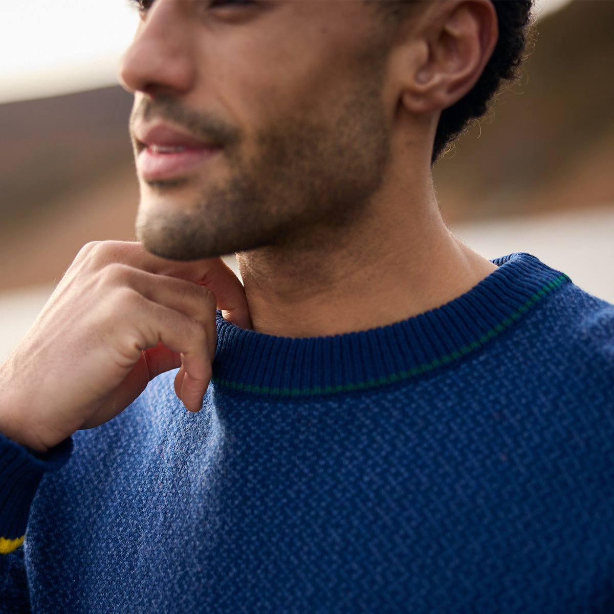 A man in a textured blue knit sweater with striped collar trim rests his hand on his chin.