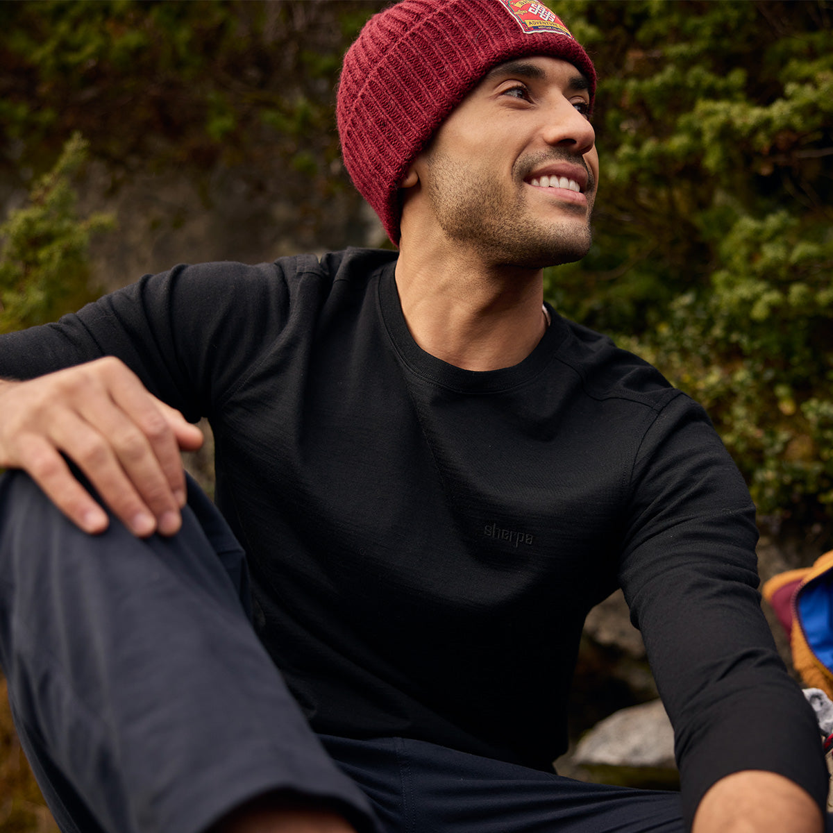 A man in a black performance shirt and burgundy knit beanie sits outdoors by green foliage.
