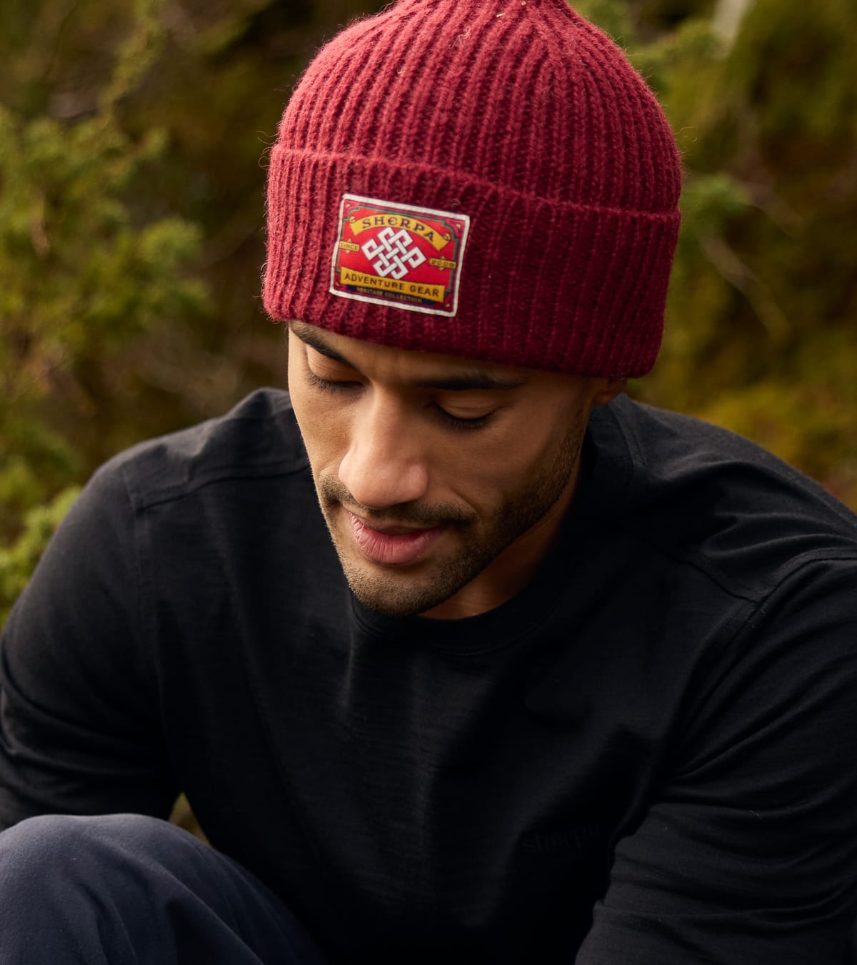 A close-up of a man looking down while wearing a deep red ribbed beanie with a Sherpa logo patch.