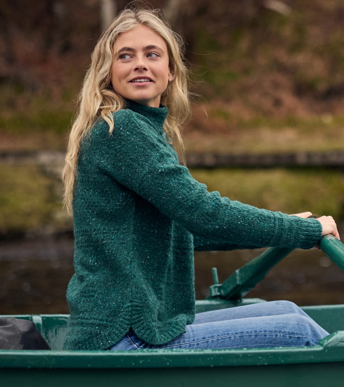 A woman wearing a dark green high-neck knit sweater rowing a boat on a lake.