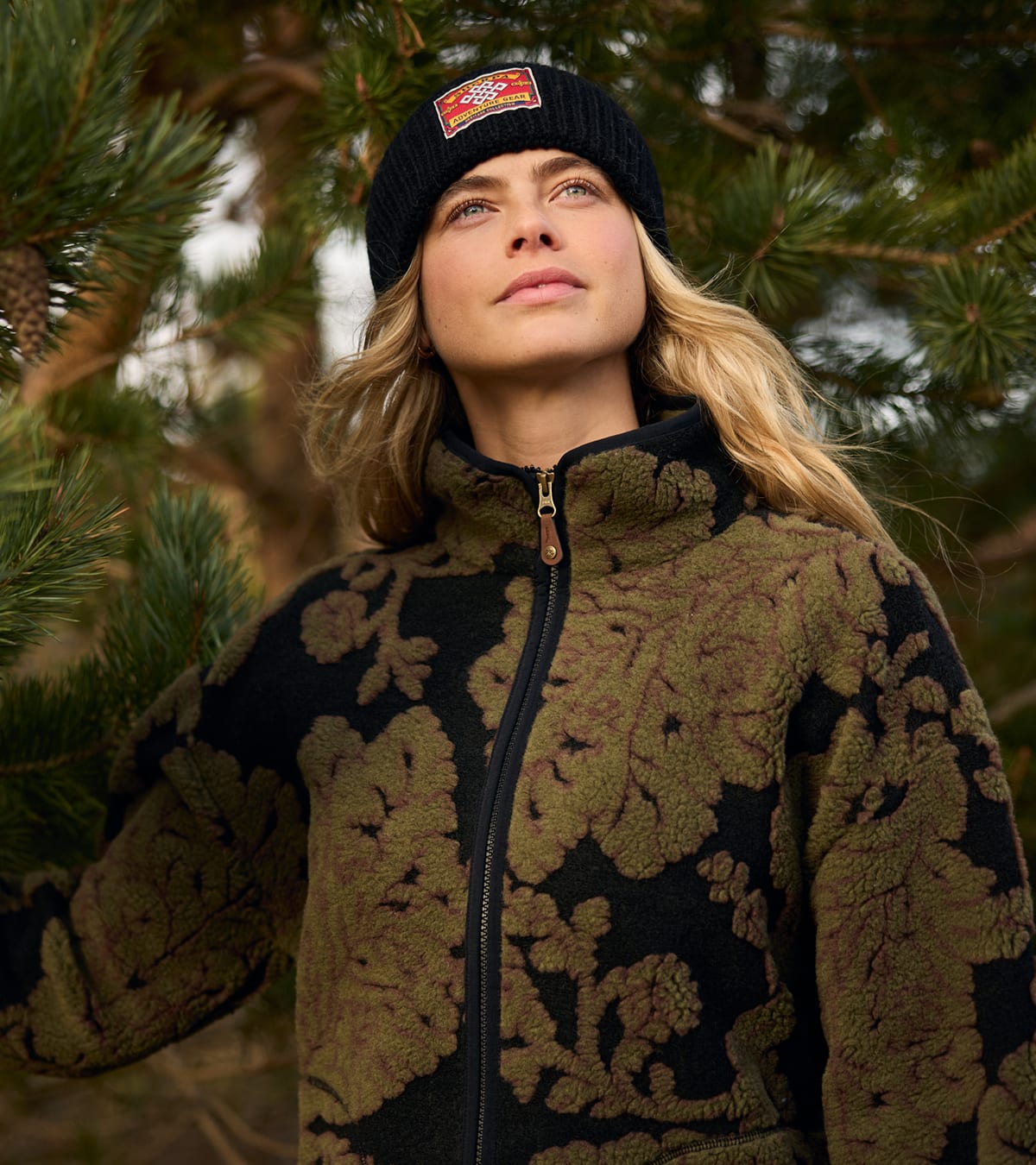 A woman wearing a black and olive green fleece jacket and a black ribbed beanie looking up.