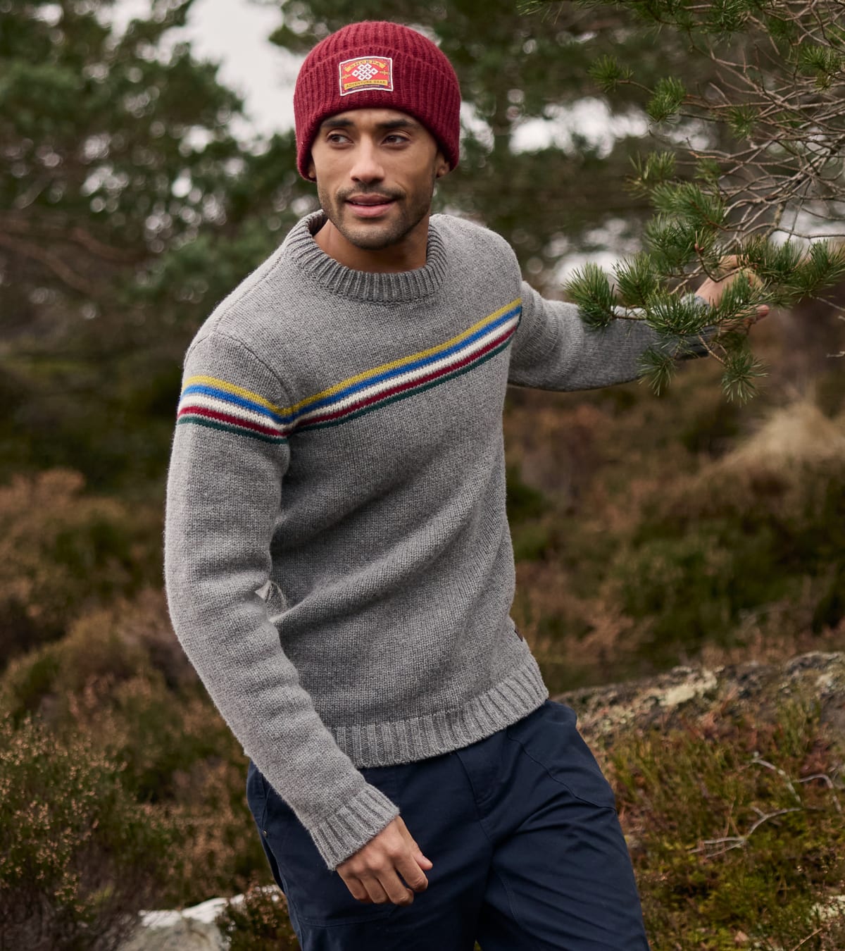 A man wearing a gray knit sweater with a colorful striped yoke, a maroon beanie, and navy trousers.