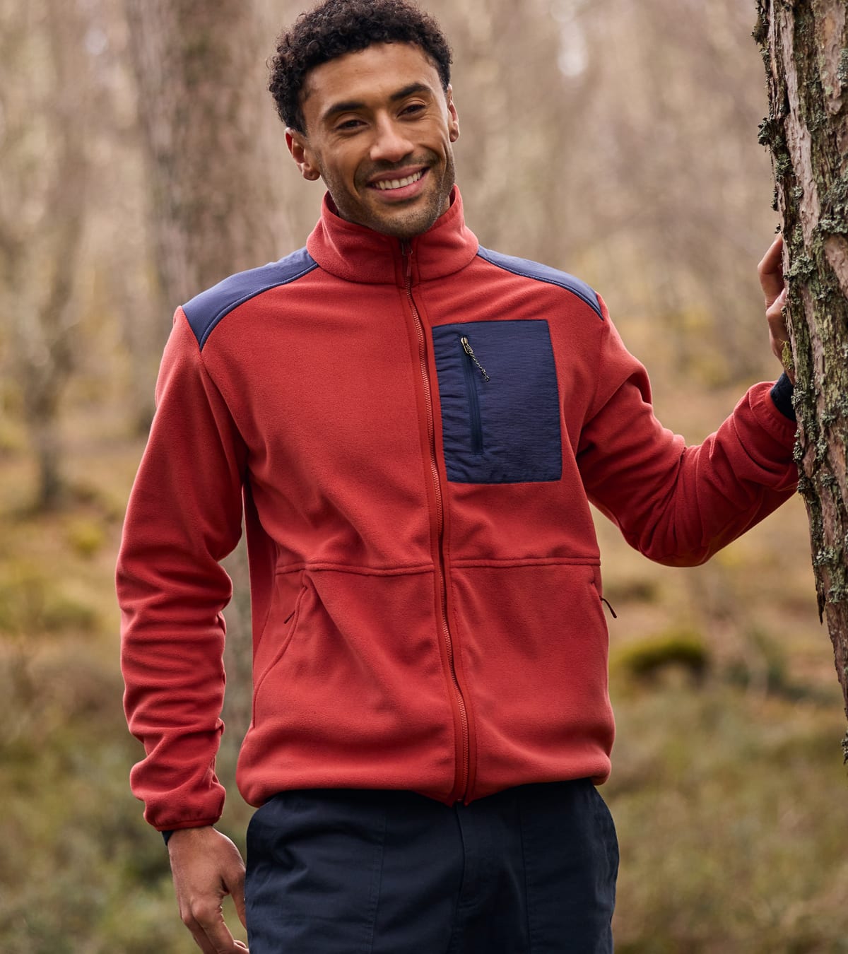 A man wearing a red and navy fleece jacket stands outdoors in a wooded setting