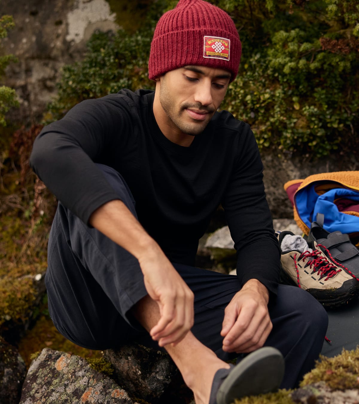 A man sitting outdoors on rocks wearing a burgundy knit beanie.