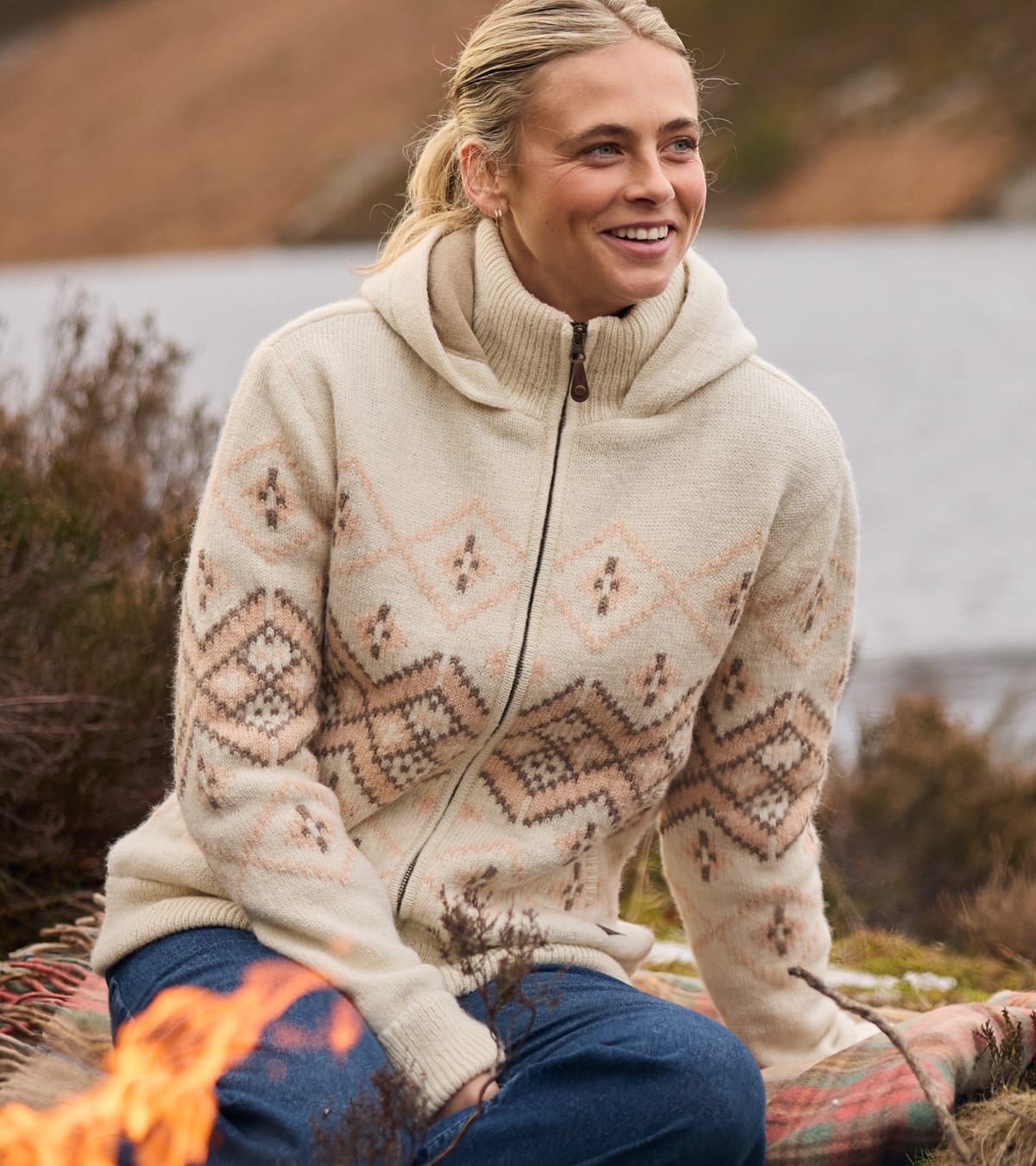 A woman sitting outdoors by a campfire wearing a cream-colored knit zip-up hoodie.