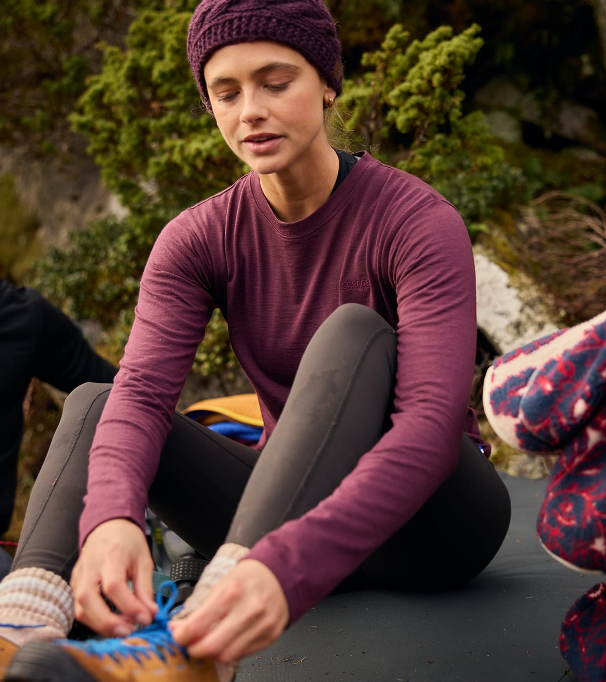A woman sitting outdoors on a dark mat, wearing a burgundy long-sleeve performance top.