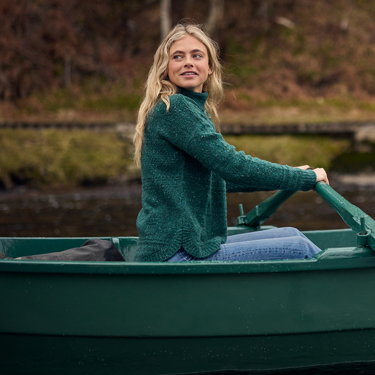 Woman sitting in a green rowboat wearing a dark green knit turtleneck and blue jeans.