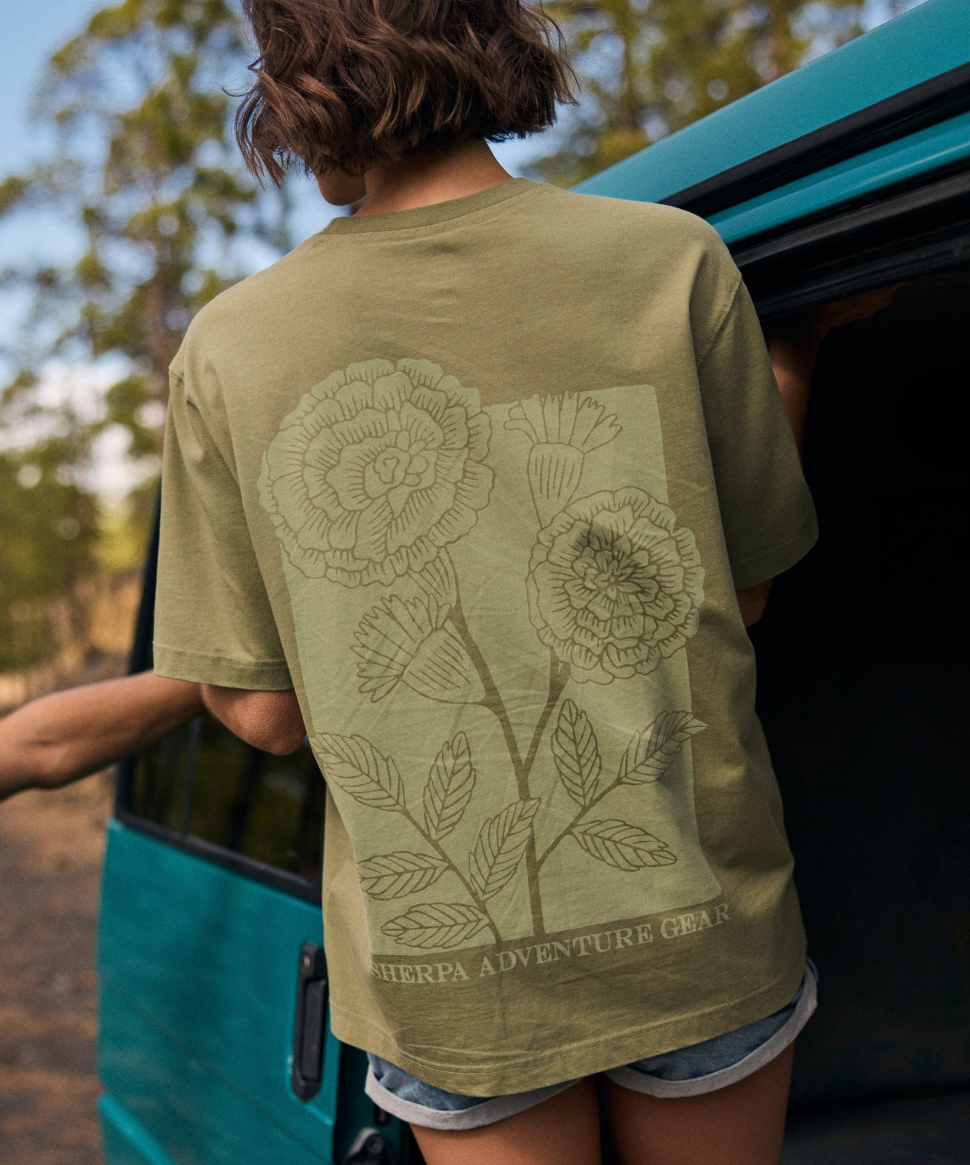 A woman wearing a green t-shirt with floral design and 'Sherpa Adventure Gear' text, standing next to a teal campervan.