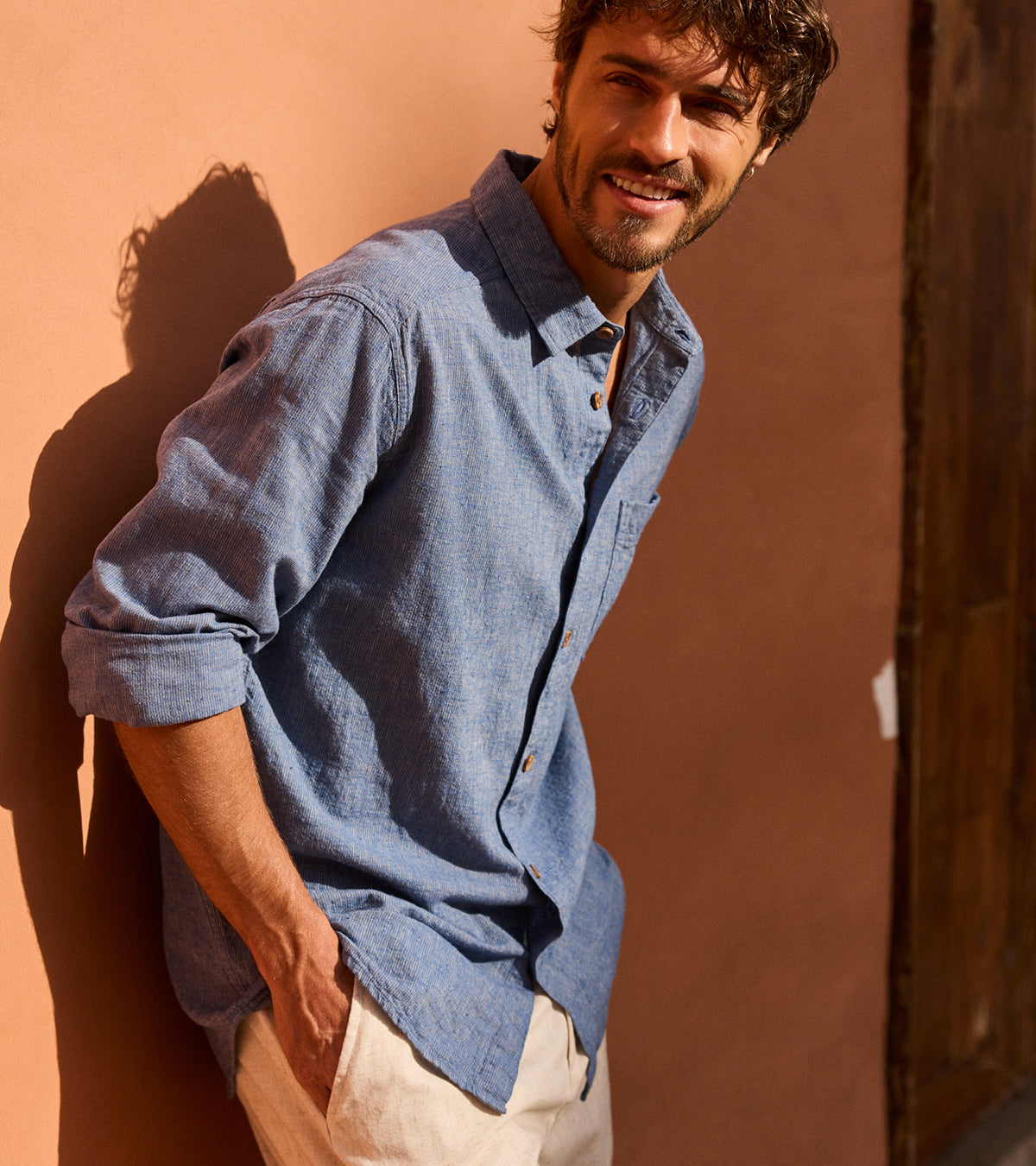 Man wearing a blue, long sleeve shirt with the sleeved rolled up against a brown wall. He is looking to his right smiling.