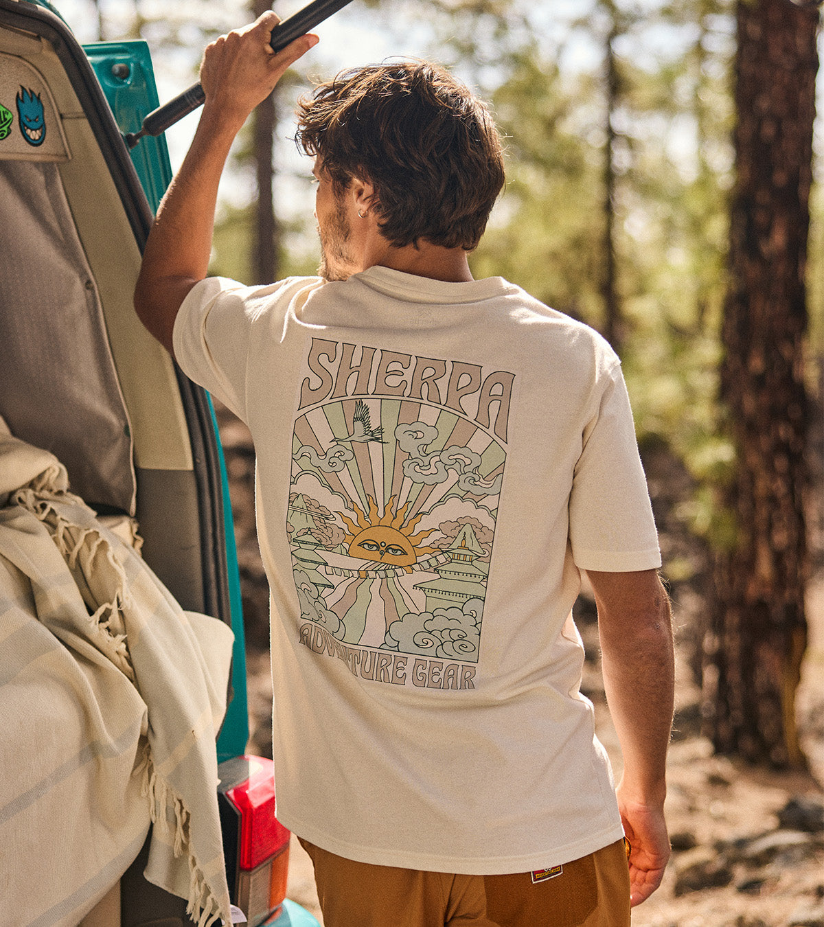 Person wearing a 'Sherpa' t-shirt with a graphic design in a forest setting. The graphic depicts a stylised Nepalese sunset with rays of light, clouds and temples.