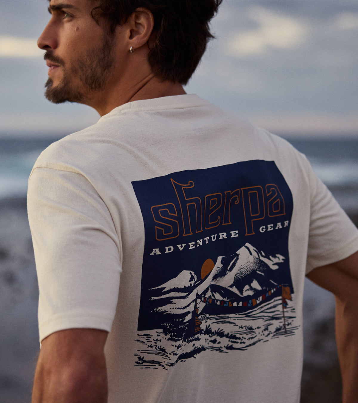 Man wearing a white t-shirt with 'Sherpa Adventure Gear' graphic depicting a mountain sunset and prayer flags in Nepal. The man is standing on a beach, looking out towards the sea.