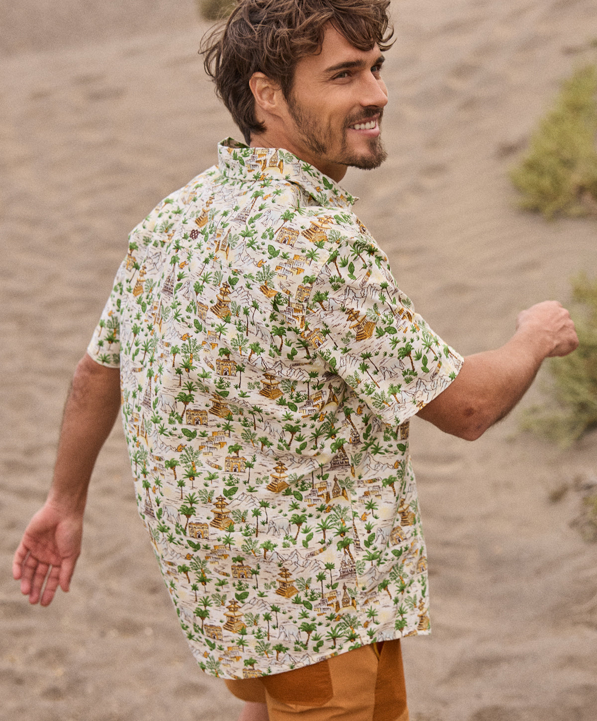 Man wearing a patterned shirt standing in a desert-like environment. The patterned shirt shows palm trees and Nepali temples.