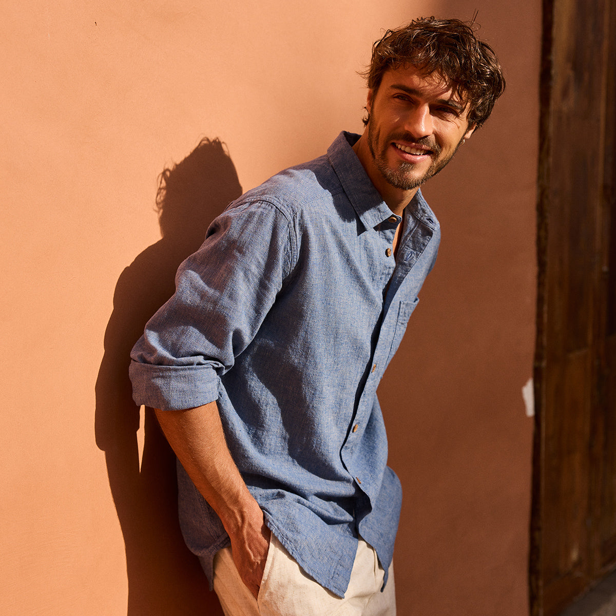 Man with curly brown hair wearing a blue shirt against a warm-toned wall.