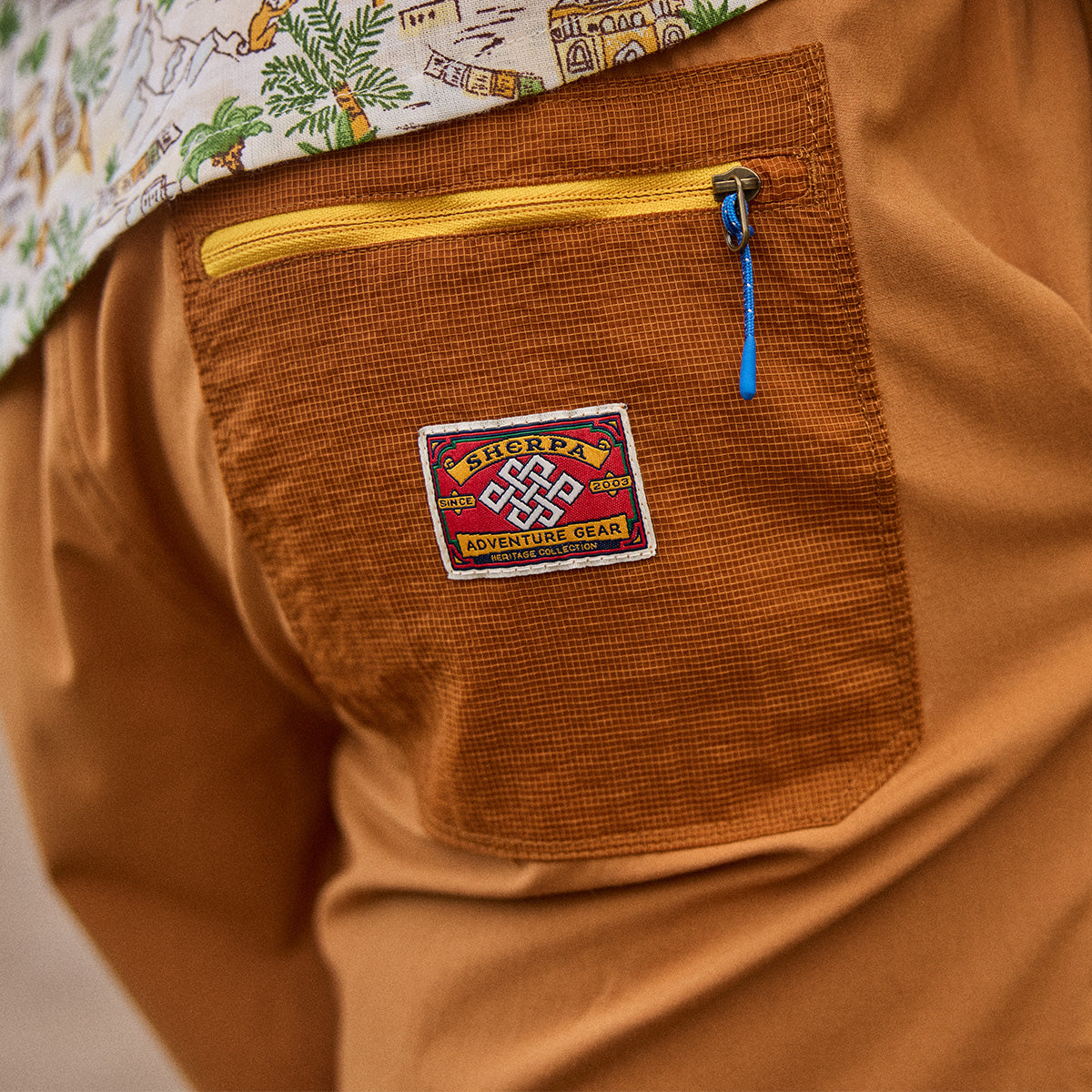 Close-up of  mustard yellow shorts with a patch and zipper pocket, featuring a "Sherpa Adventure Gear" patch.