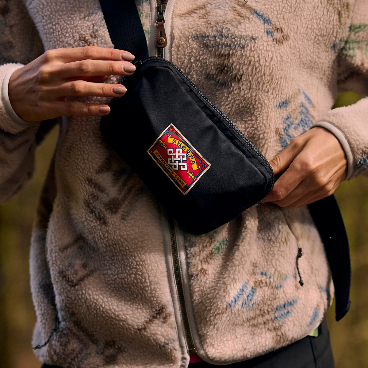 A close up of a woman wearing a patterned fleece and holding a black bag with a colorful "Sherpa Adventure Gear patch" on the front.