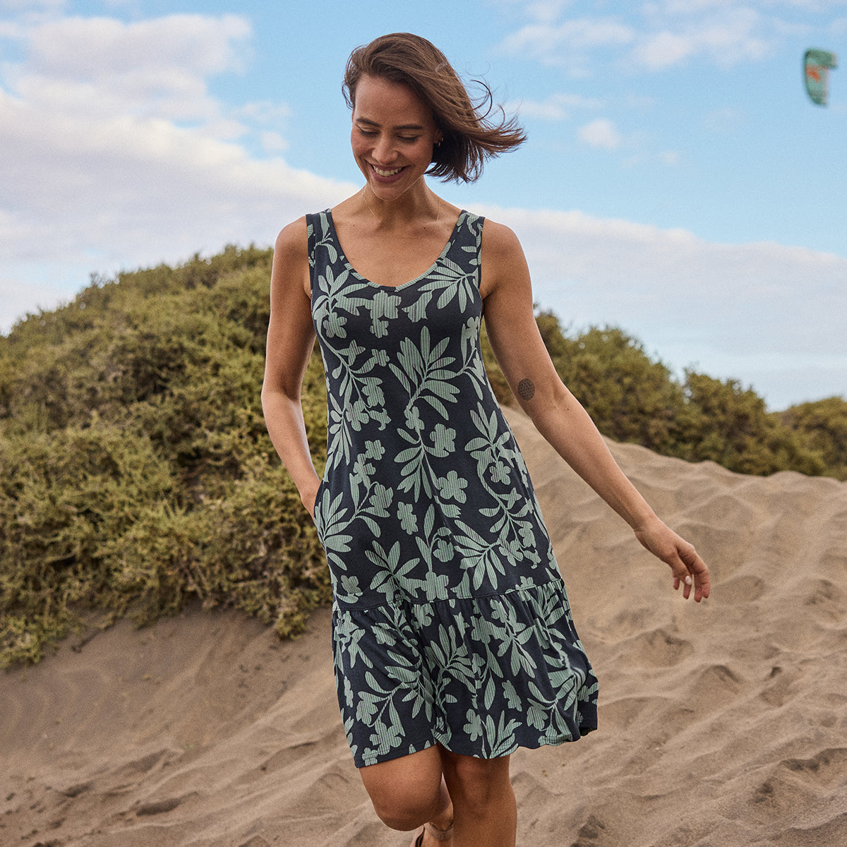 Woman in a floral dress walking on a sandy beach with greenery and blue sky in the background