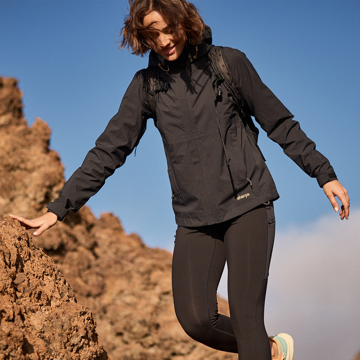 Person in black waterproof jacket and pants hiking on rocky terrain with a clear blue sky.